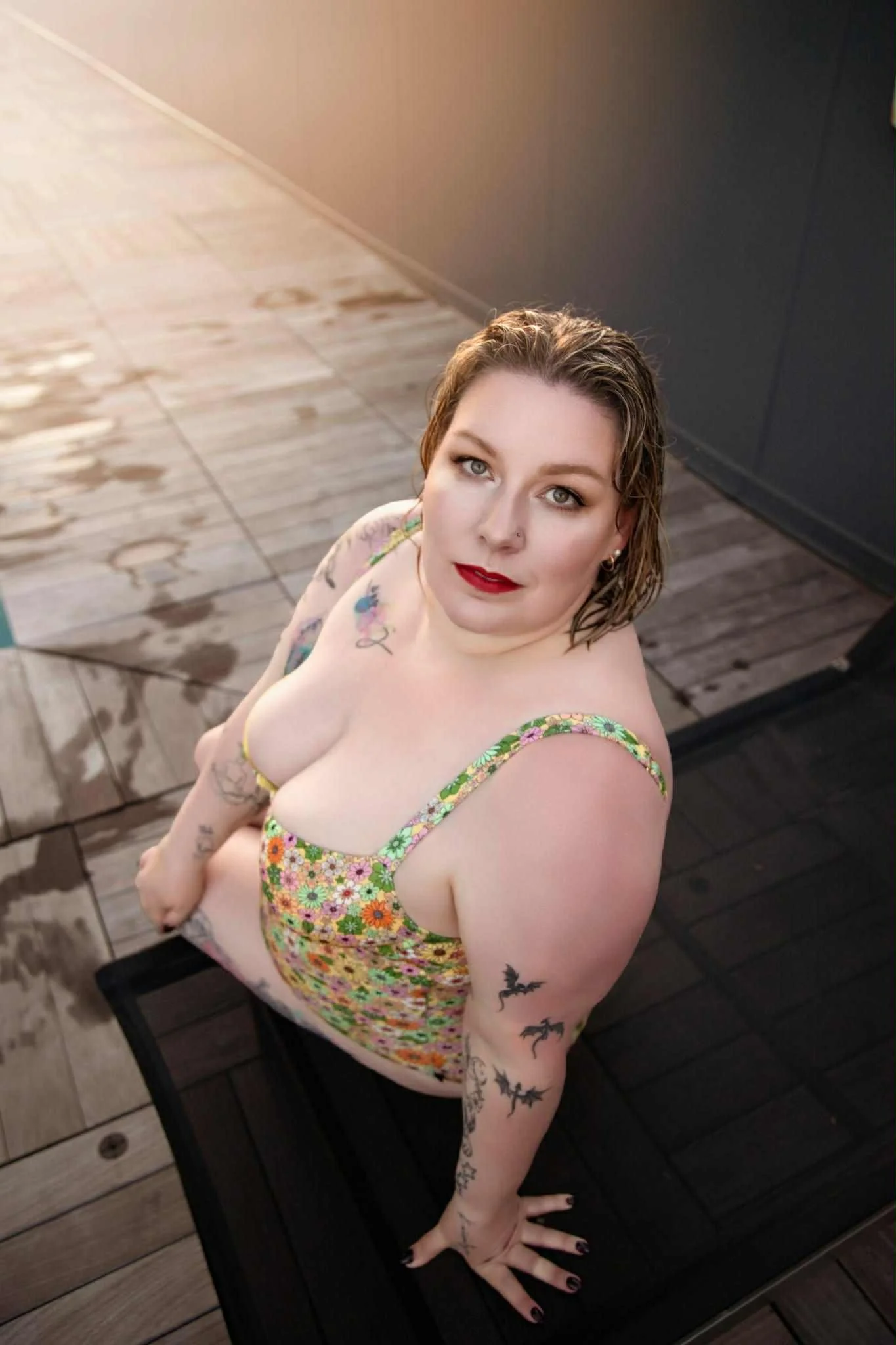 A woman with short wet hair, wearing a colorful floral dress, is kneeling on a black surface outdoors, looking up at the camera with a confident expression. She has tattoos on her arms and shoulders, and is wearing red lipstick and makeup.