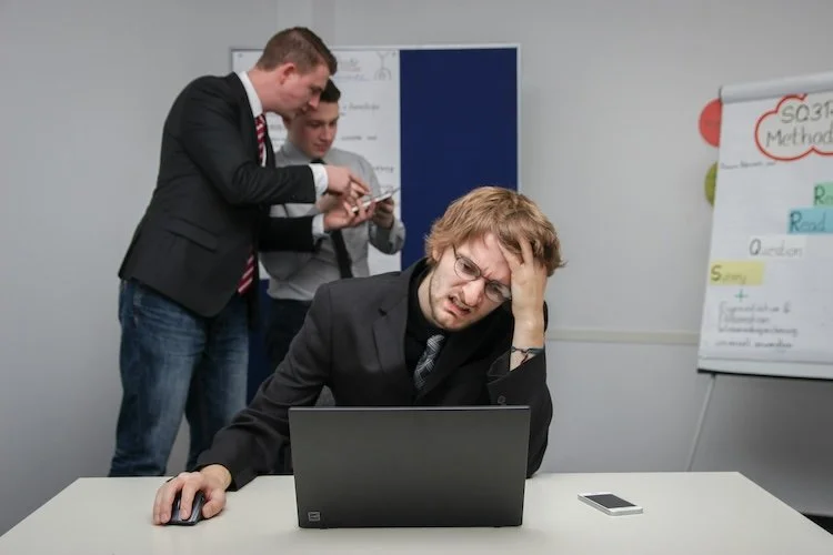 Man sitting with a stressed look on his face, staring at a laptop, with two men behind him staring at another device.  They need to take back control at work.