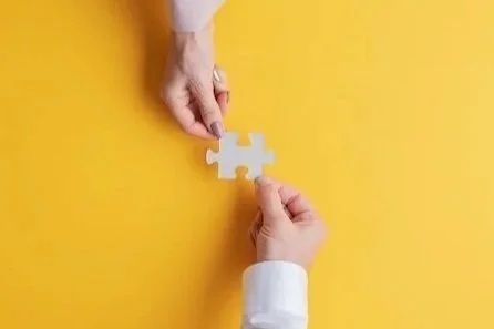 Two hands holding a jigsaw piece over a yellow background.