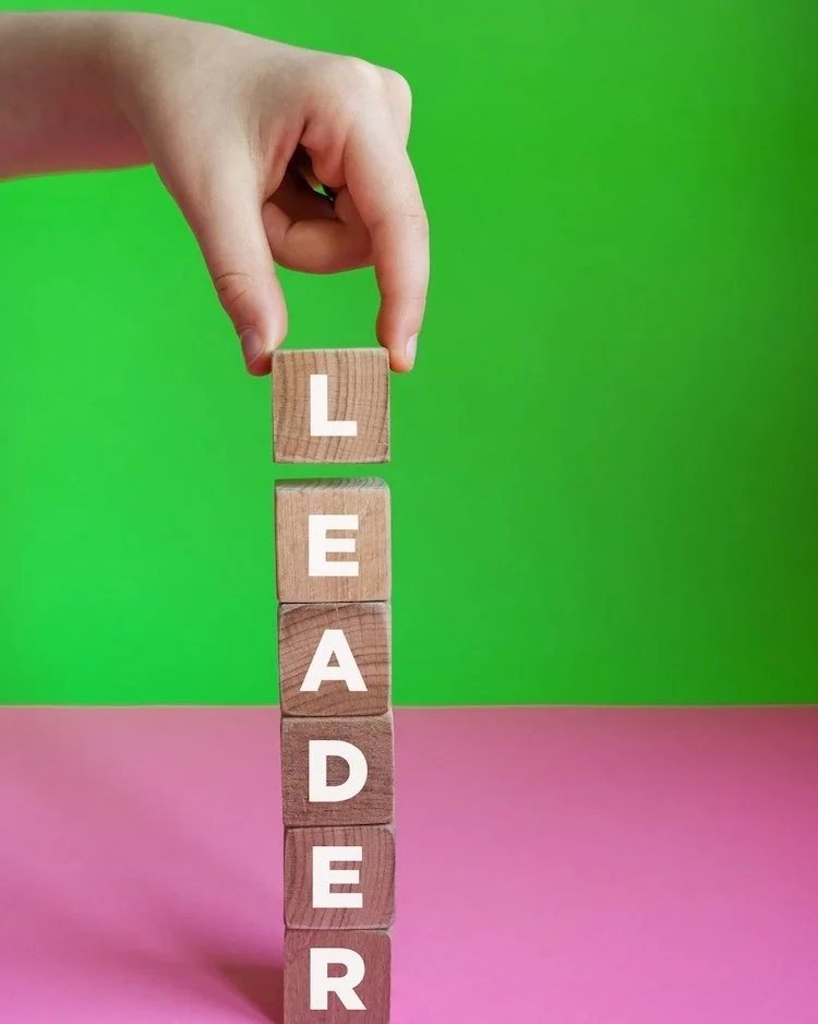 Woodlen building blocks with the word 'leader' on them. This image is on a pink desk and green walls and represents steady leadership gained from the Not winging It leadership Club.