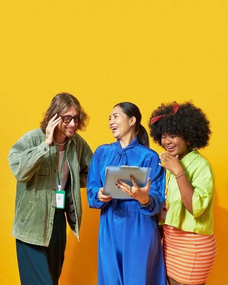 A mand and two women in bright coloured clothes, standing in front of a yellow background, looking at a list in the hands of the middle person. It is to represent why you would join the Not Winging It Leadership Club as a founding member.