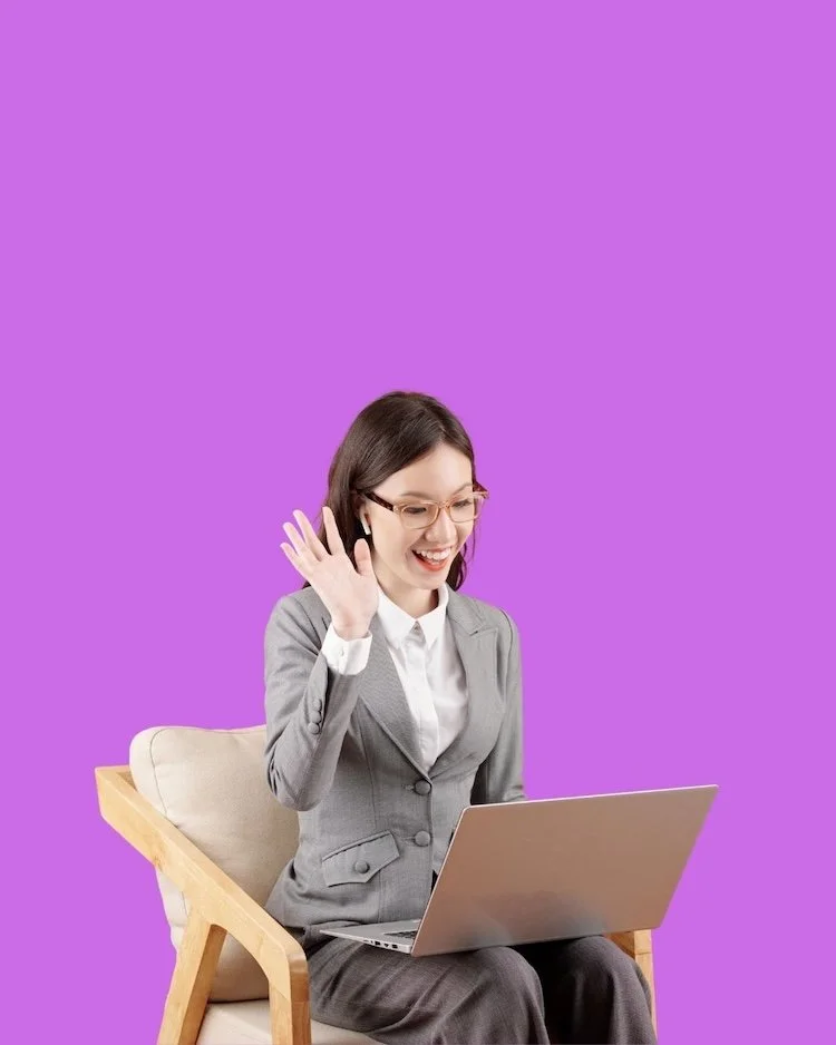 Image of long haired woman in suit, waving at her laptop, whilst sitting on a seat, in front of a purple background. To represent the monthly live calls at The Not winging It Leadership Club.