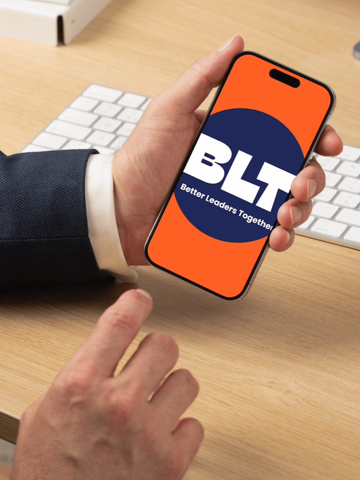Image of man in a suite at his desk with keyboard,  holding a mobile phone with the app BLT on it.