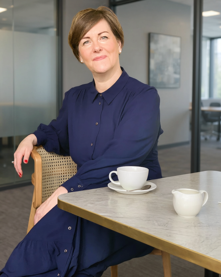 Sharon Smith of Own Your Leadership is wearing a blue dress with long sleeves and buttons up the front. She is sitting on a wicker chair, at a table with a cup of tea in front of her.