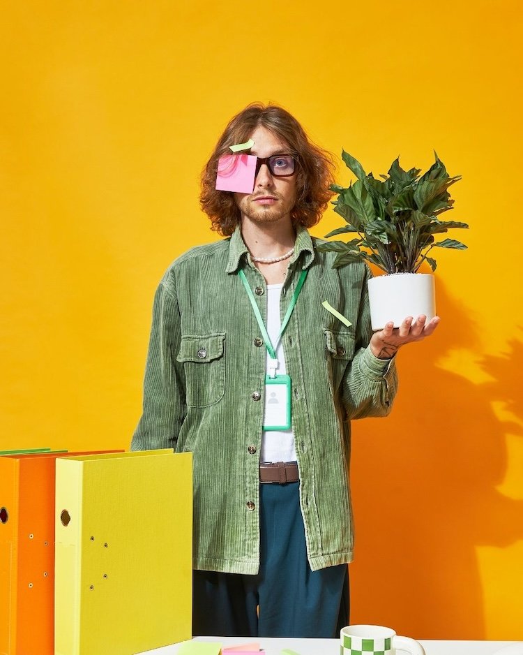Man with long brown hair standing in front of a desk holding a plant and has sticky notes stuck to his glasses. He looks unhappy. The background is orange and there are two brightly coloured files on the desk in front of him.