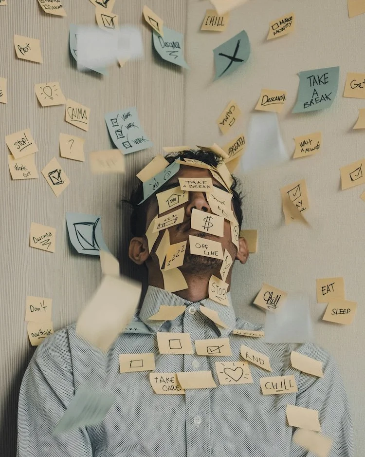 man hiding in a corner with lots of post it notes on his face - he is clearly overloaded at work.