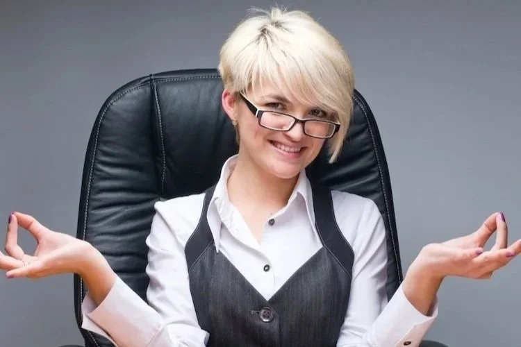 Image of a blonde haired woman with glasses sitting at a board table wearing a white shirt and grey waistcoat smiling to represent that she is the right type of person for the Not Winging It Leadership Club.