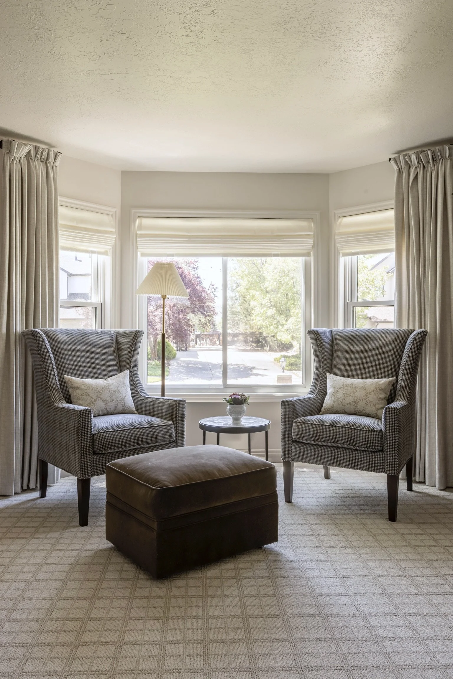 Living room with two armchairs, a small table with a flower vase, a brown ottoman, a floor lamp, and large windows with curtains and shades, overlooking a street with trees.