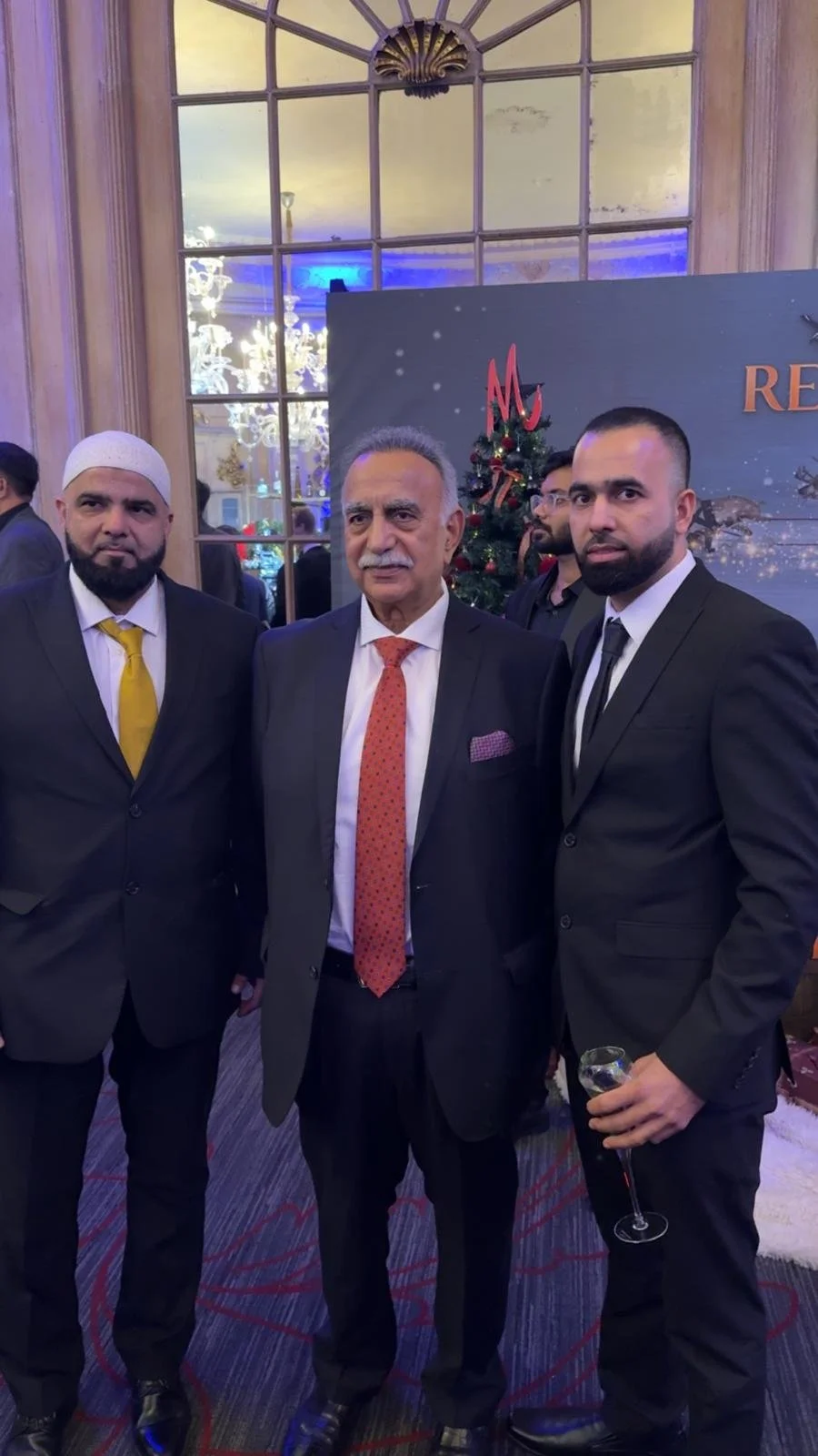 Three men in formal suits posing for a photo at a holiday event, with a Christmas tree and festive decorations in the background, and a chandelier reflected in the mirror behind them.