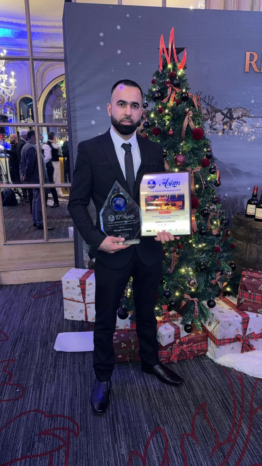 Man in formal suit holding an award and a framed certificate standing next to a decorated Christmas tree with wrapped presents underneath.