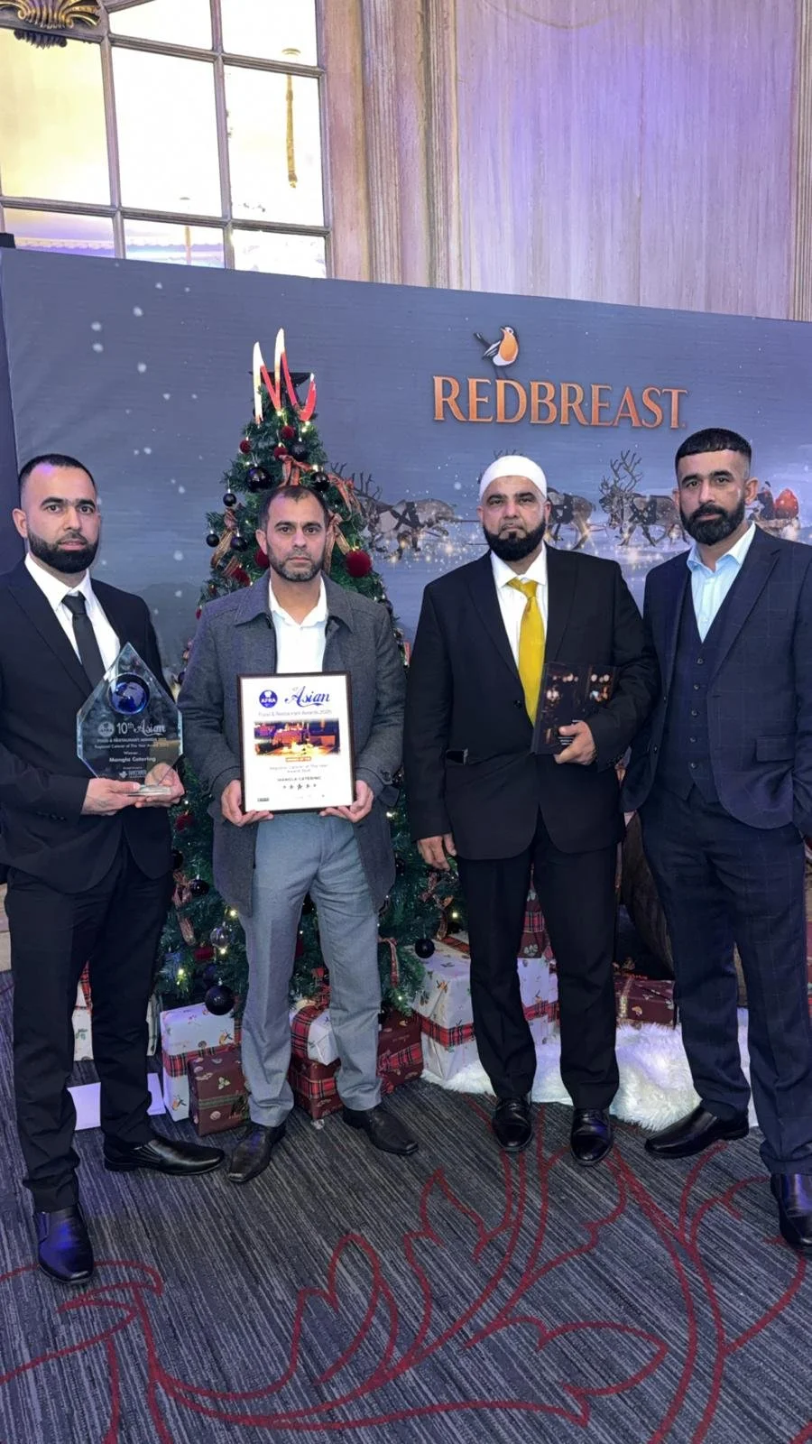 Four men dressed in formal suits and ties standing in front of a decorated Christmas tree with wrapped presents underneath. They are holding awards or certificates. Behind them is a backdrop with the word 'REDBREAST' and a bird logo, suggesting an ev