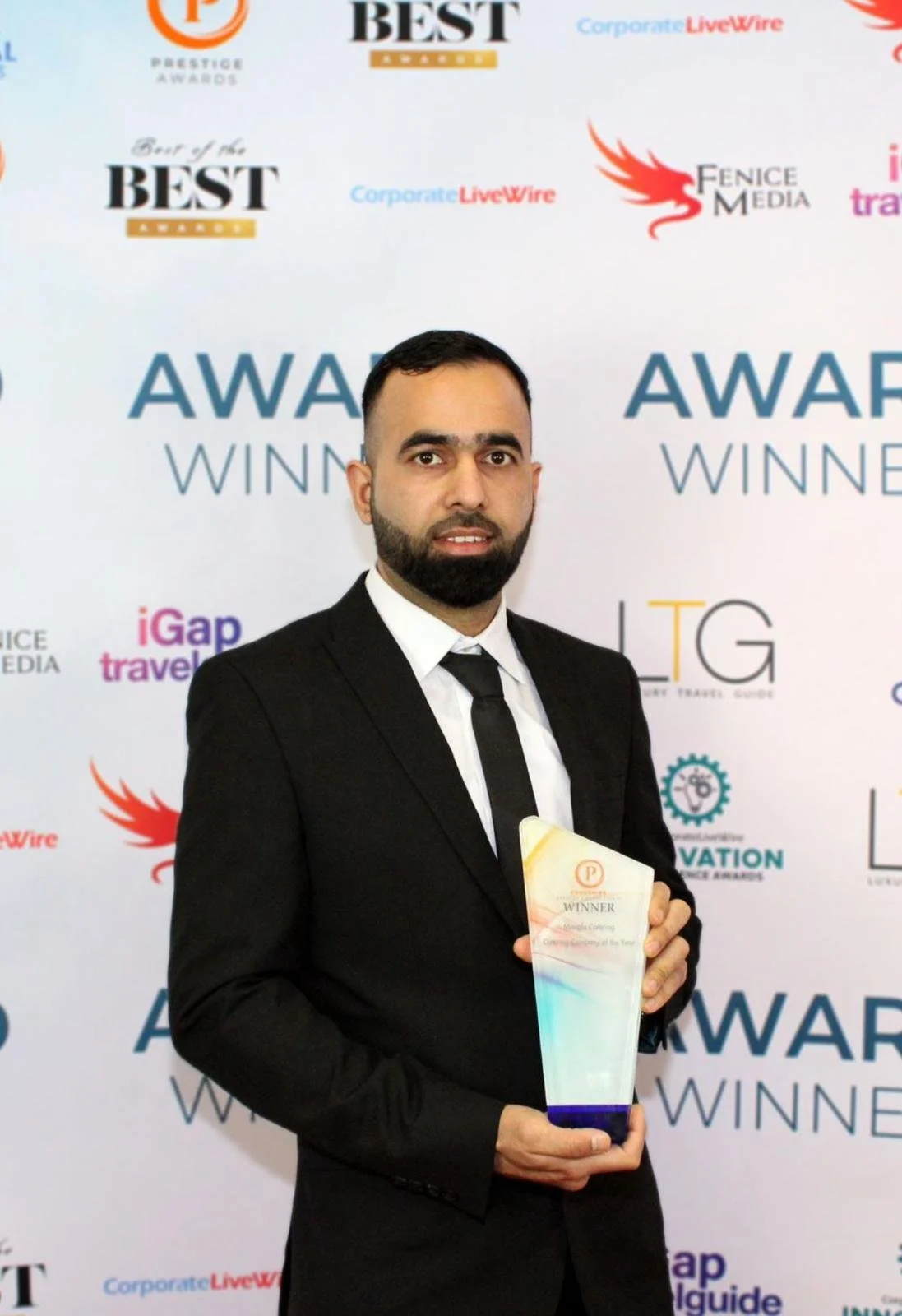 A man in a black suit and tie holding an award trophy at an award ceremony. The backdrop features multiple logos and the words 'AWARD WINNER'.