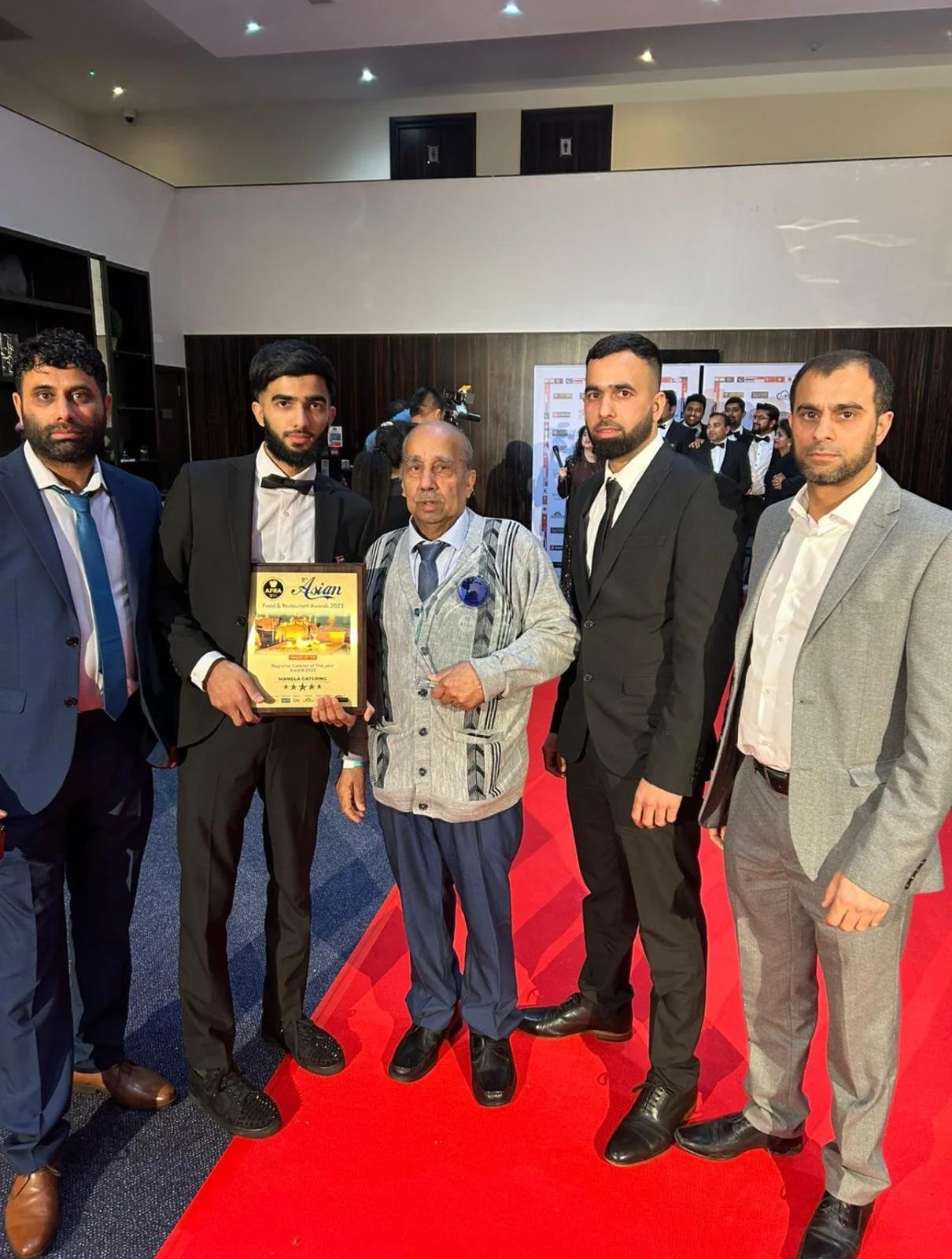 A group of six men in formal attire standing on a red carpet at an awards event, with one holding a framed award plaque.