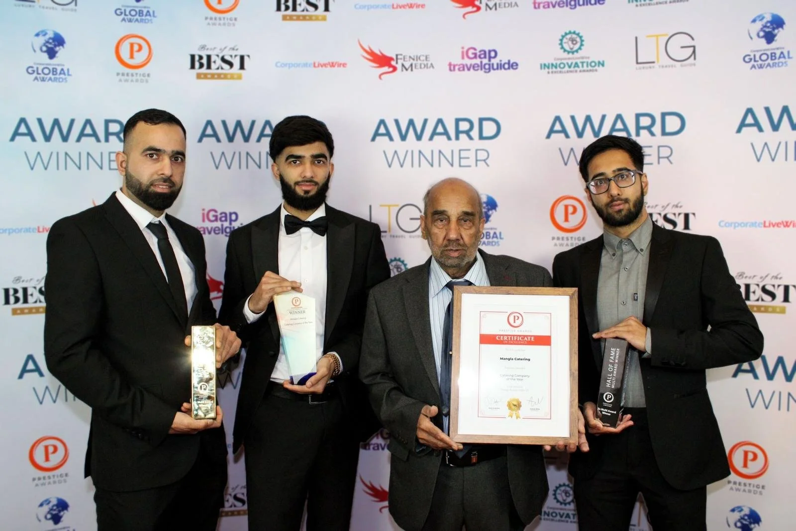Four men standing in front of a backdrop with awards logos, holding trophies and a framed certificate. The group appears to be celebrating an award win.