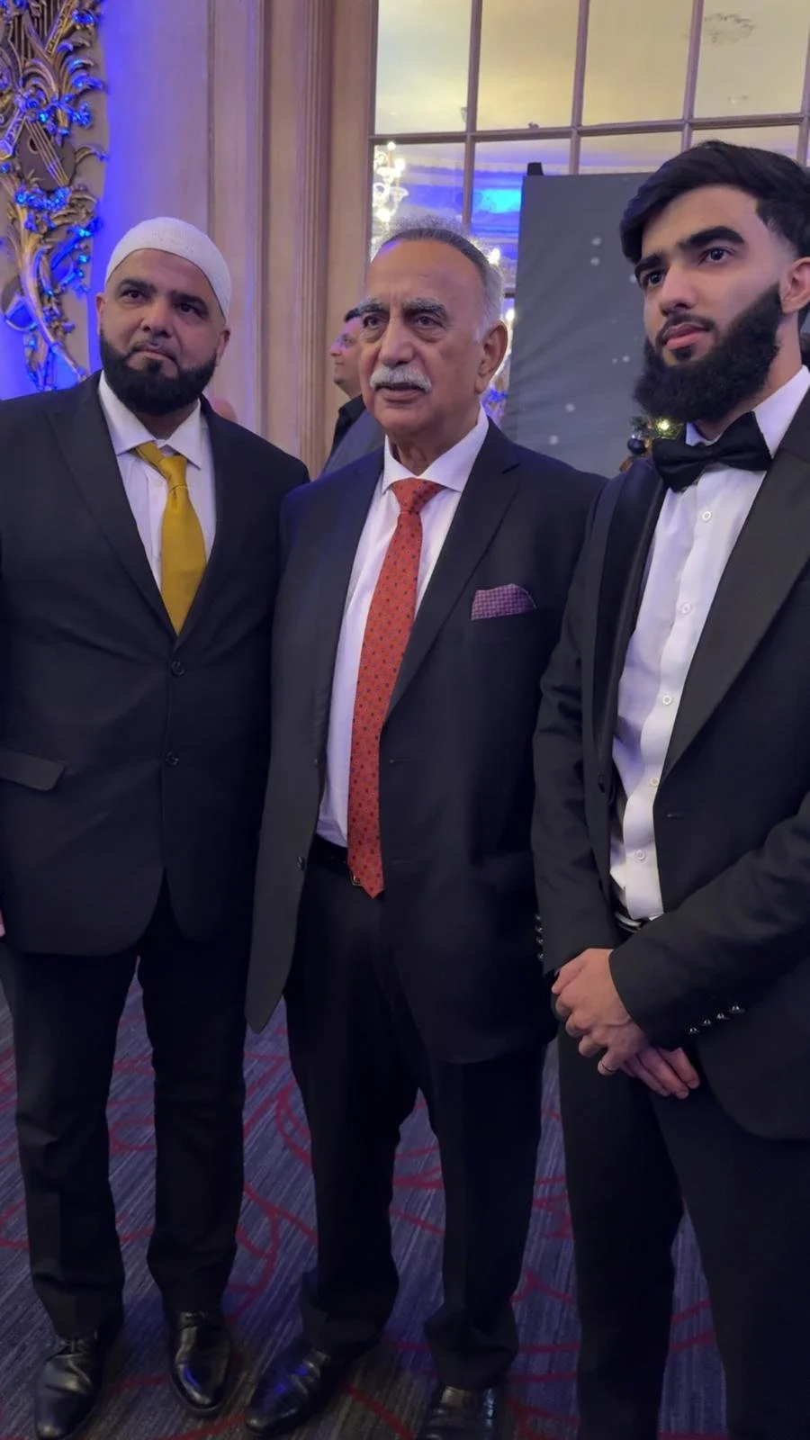 Three men in suits at a formal event, standing indoors near a decorated wall and window, with elegant lighting.