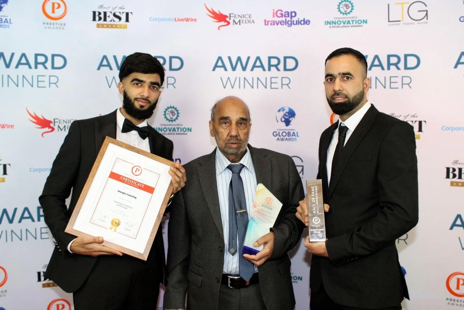 Three men in formal suits and ties standing in front of a white backdrop with award logos, holding certificates and trophies after an award ceremony.