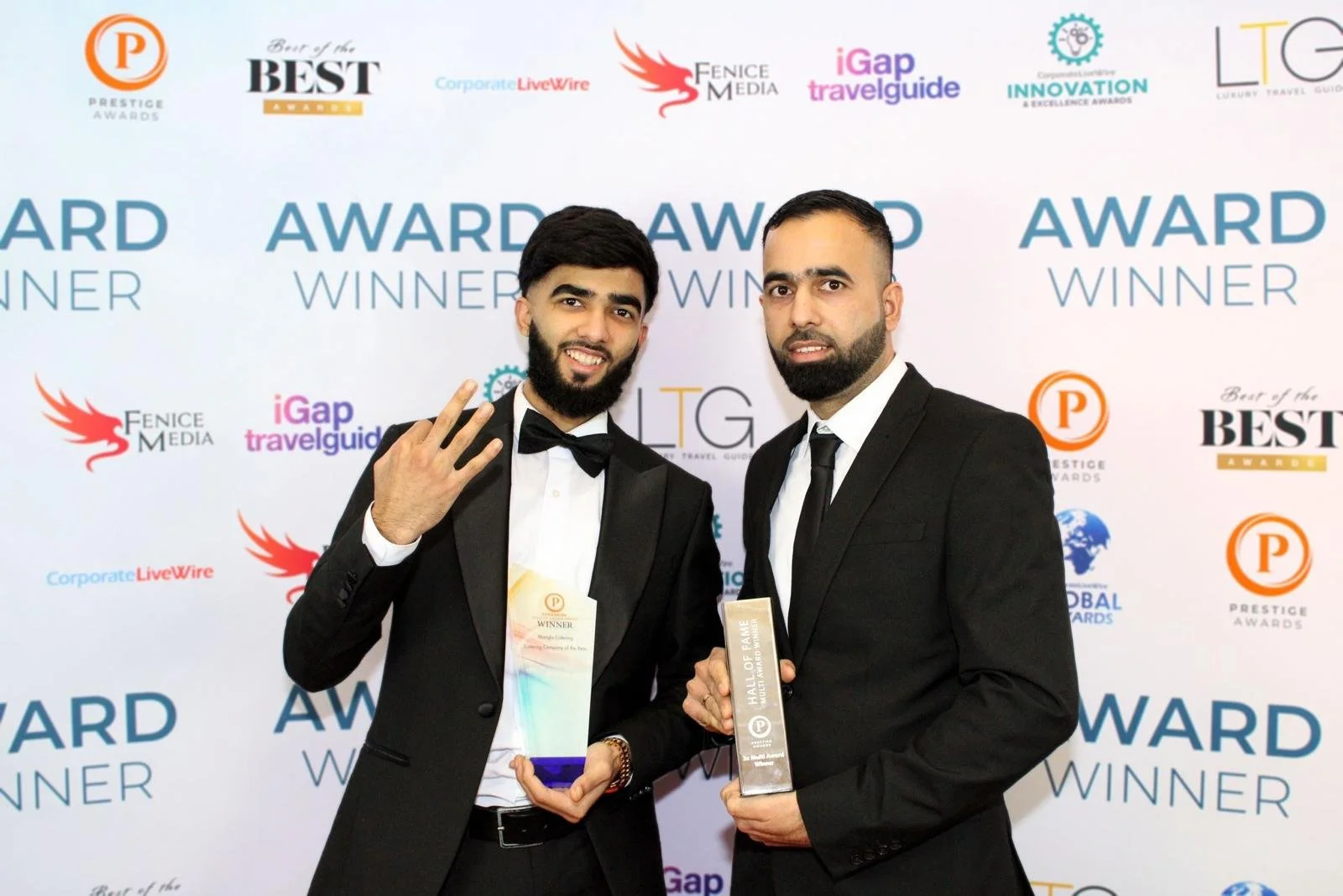 Two men in black tuxedos holding awards at an award ceremony, standing in front of a backdrop with various sponsor logos and the words 'AWARD WINNER'.