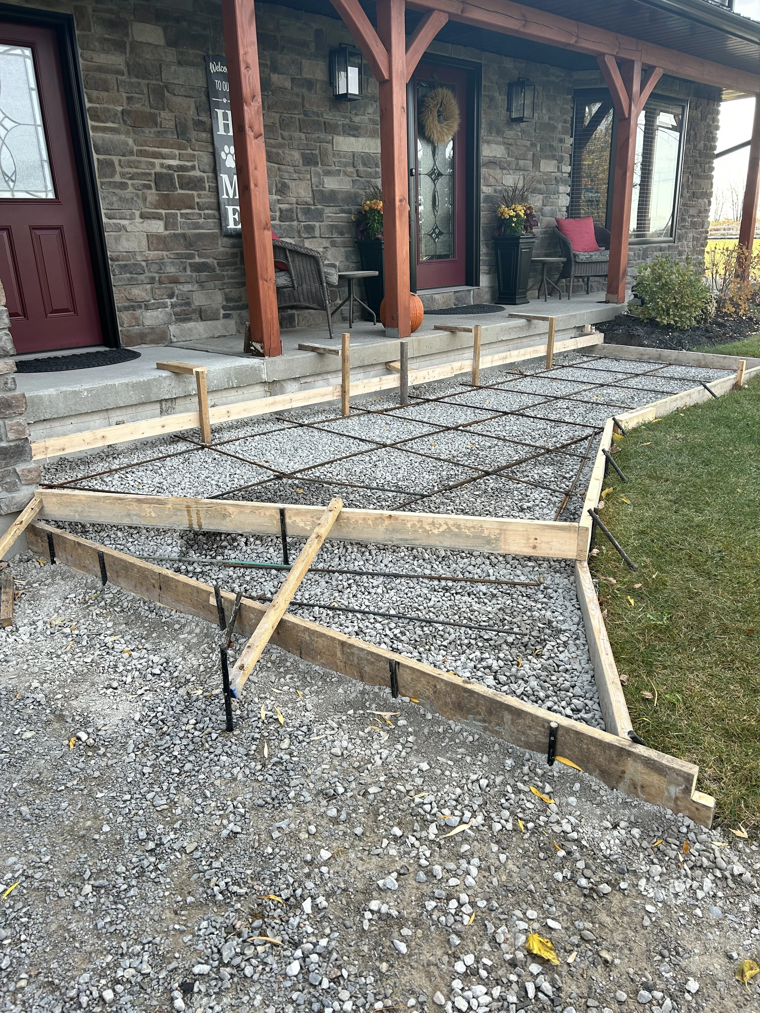 A front porch with a vegetable garden bed being prepared with wood and black mesh landscaping fabric, surrounded by gravel and a grassy lawn, in front of a brick house with decorated front door and seating area.