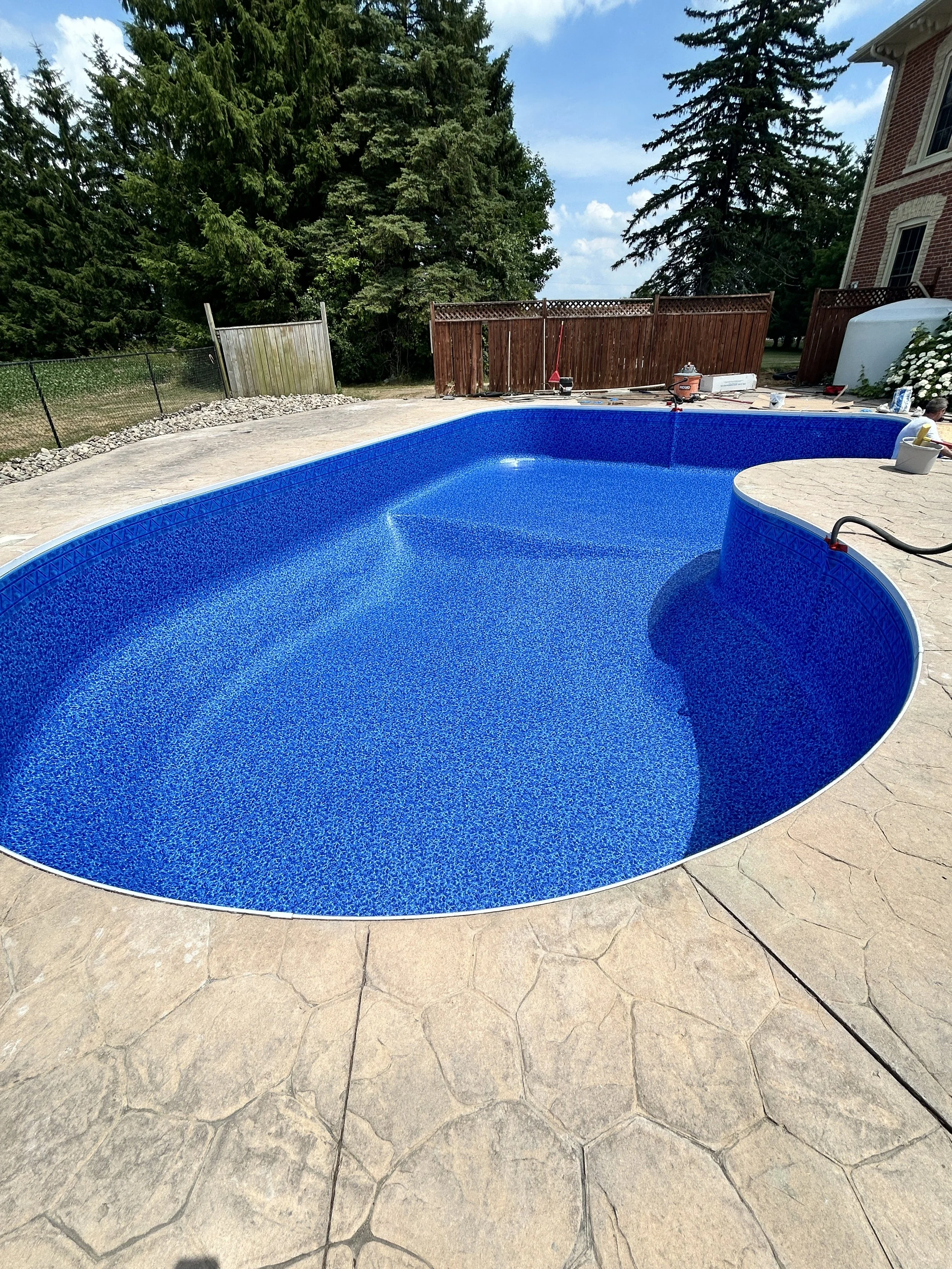 Empty in-ground swimming pool with blue lining and textured beige patio surrounding it. Fence and trees in the background, with a house partially visible on the right. Pool equipment and tools are placed on the far side of the pool.