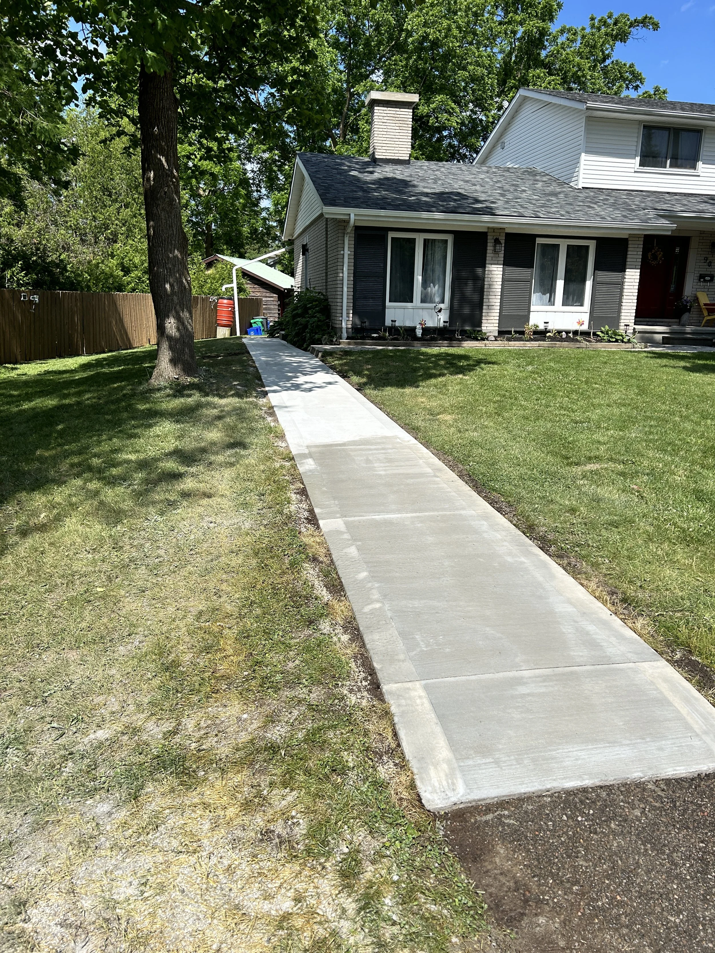 Concrete sidewalk leading up to the front of a house with a porch, surrounded by a well-kept lawn and a large tree, under a clear blue sky.