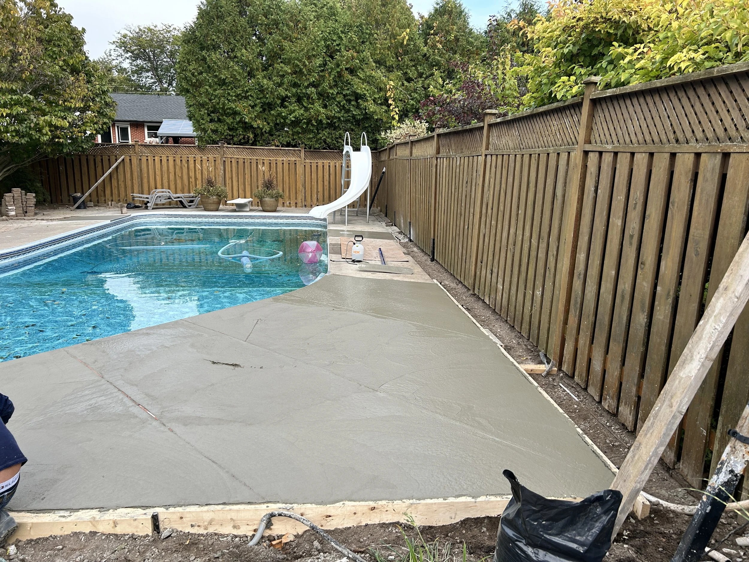 A backyard swimming pool with a new concrete slab being poured and leveled. There is a white slide at the far end, pool cleaning equipment floating in the water, and outdoor furniture including lounge chairs and potted plants along the fence.
