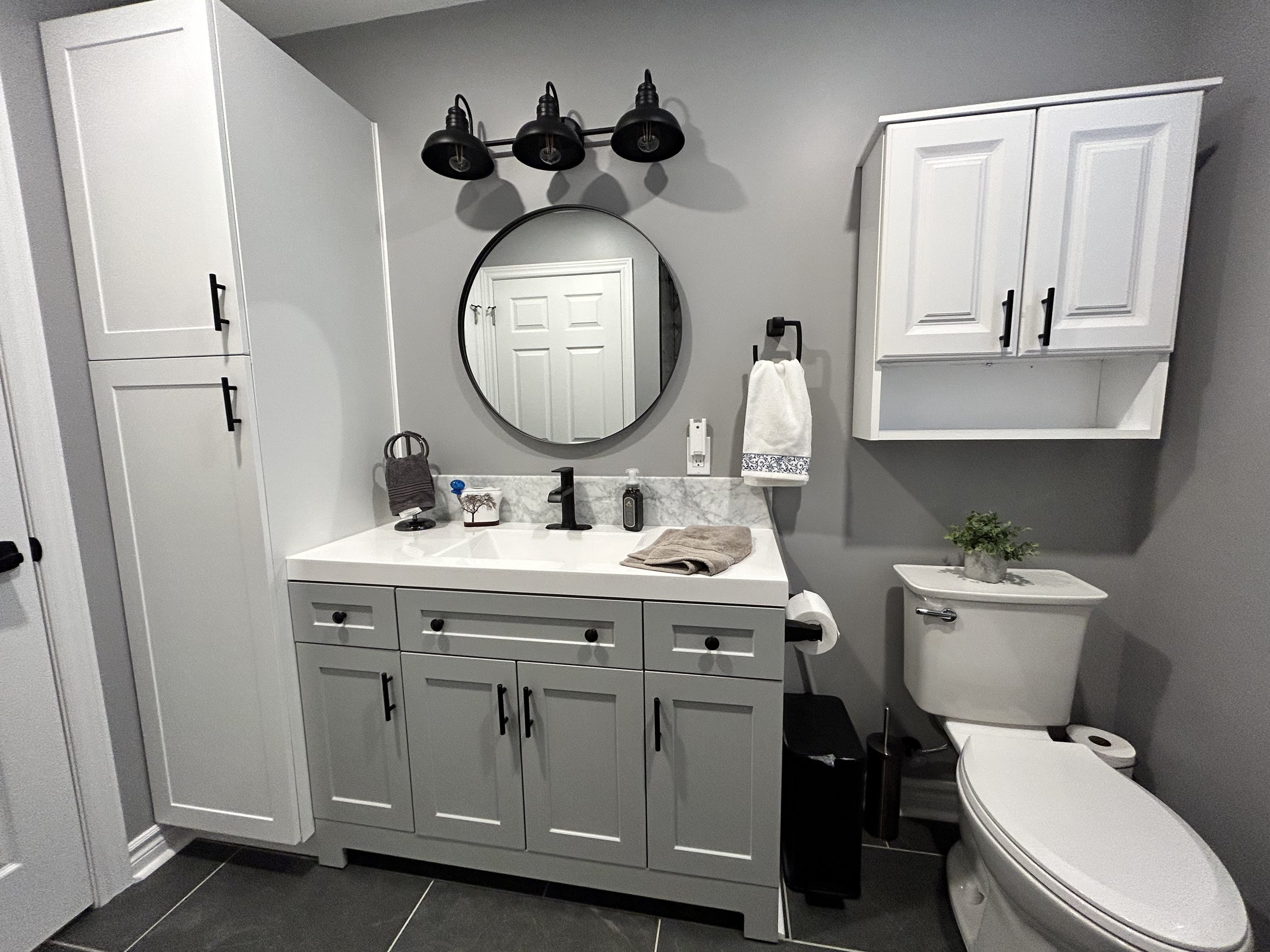 Bathroom with gray walls, white cabinetry, a black and white oval mirror, and black light fixtures. The vanity has a black faucet, soap dispenser, and a towel. There is a toilet with a small potted plant on top and a white cabinet above it. The floor