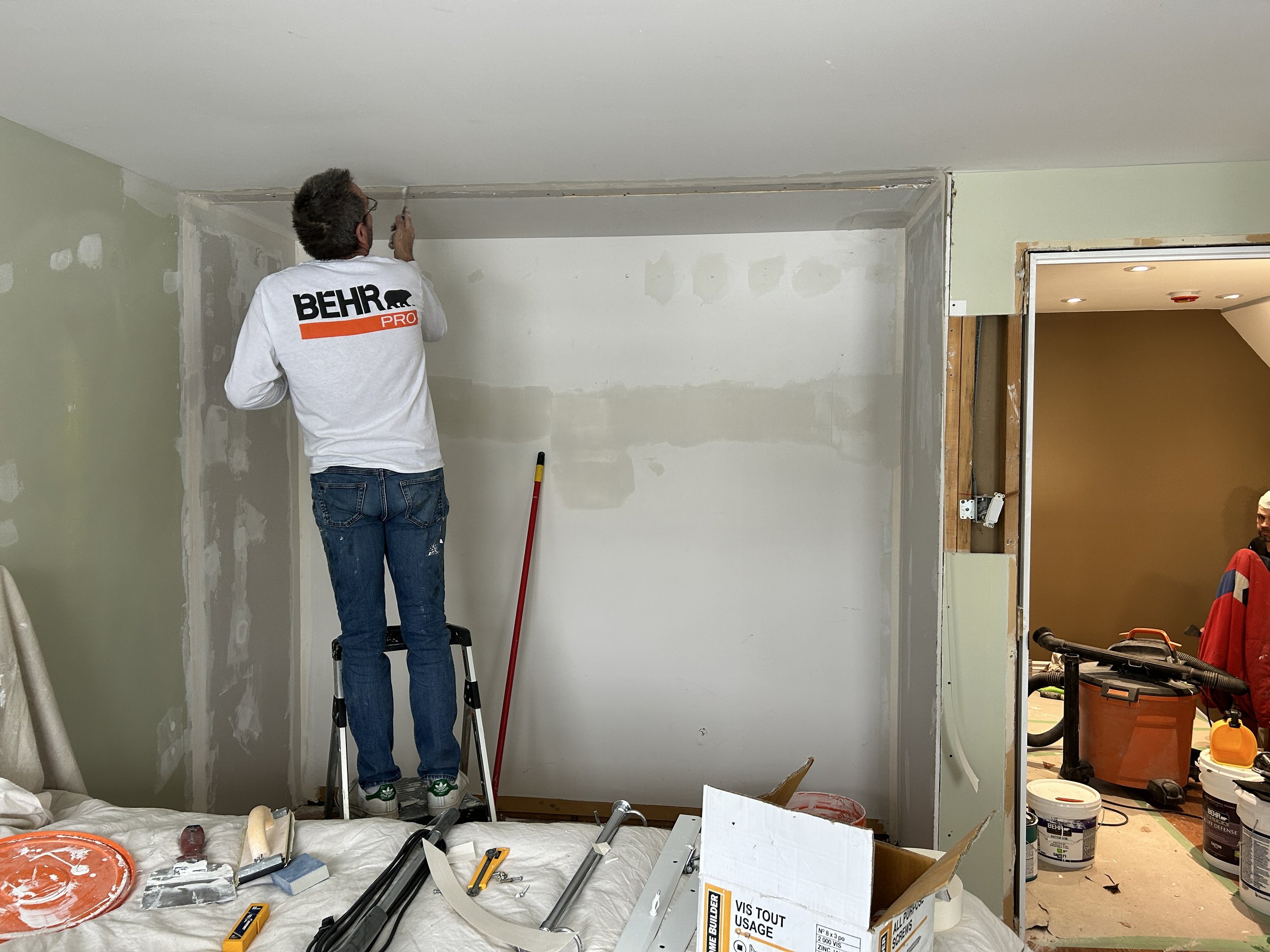 A man standing on a ladder installing molding along the ceiling in a room under renovation, with tools and supplies scattered on a covered surface in the foreground.