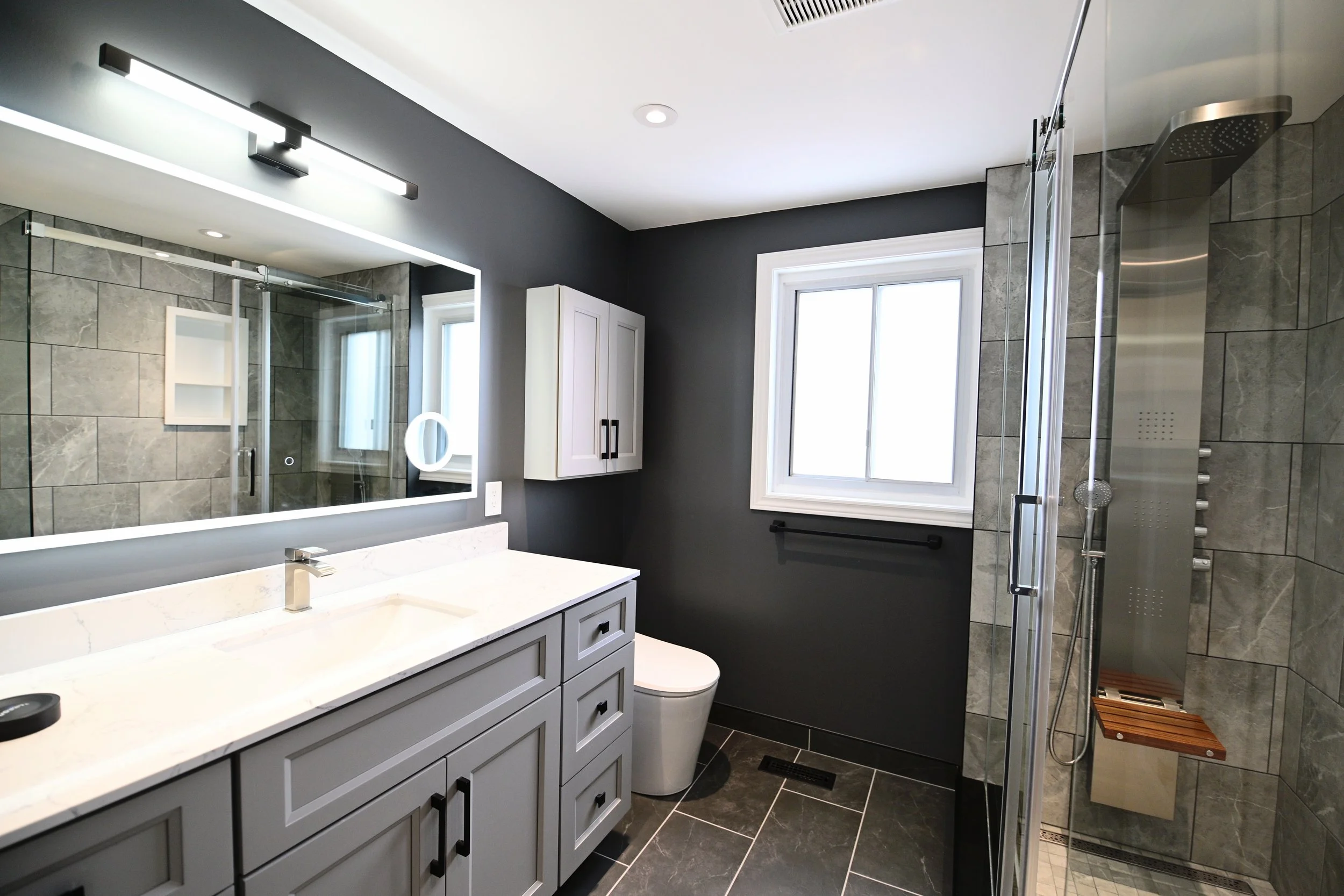Modern bathroom with gray cabinetry, white countertop, large mirror with backlight, toilet, window with black towel bar, gray tiled shower with glass door and shower panel, small wooden stool inside shower, dark gray floor tiles, and dark gray wall p