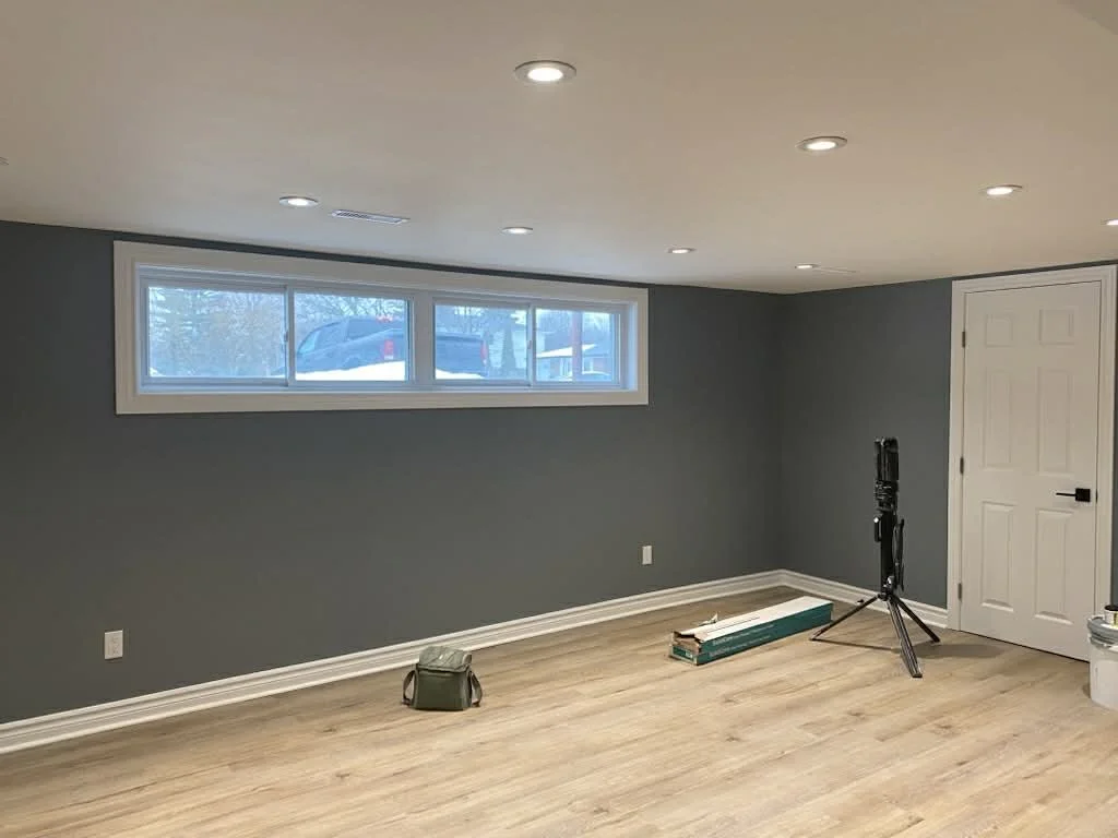 Empty finished basement with gray walls, wood flooring, a row of small windows, and ceiling lights, with some construction tools and materials near the wall.