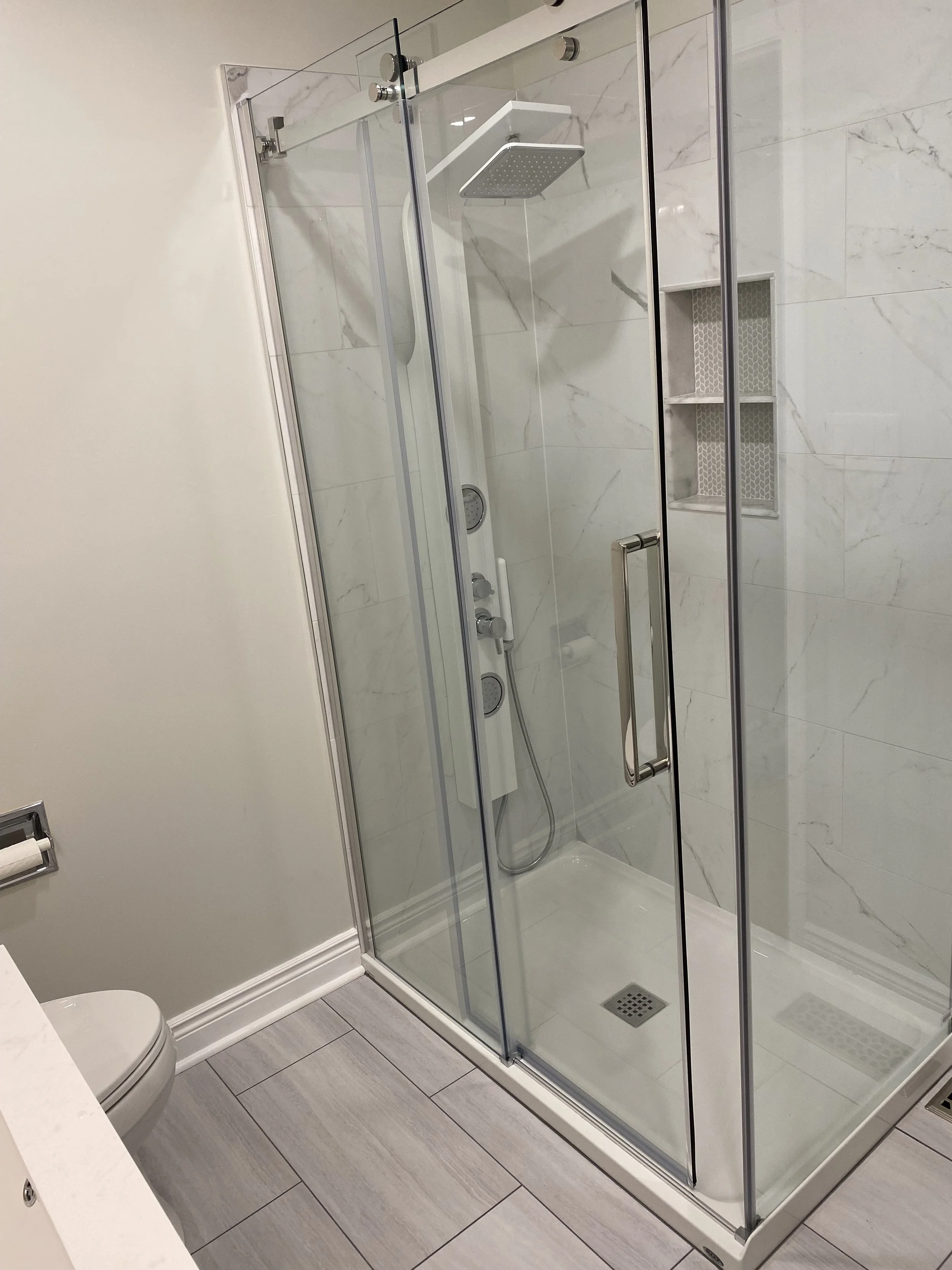 Glass shower enclosure with white marble wall tiles, built-in storage niches, shower head, and fixtures in a modern bathroom.