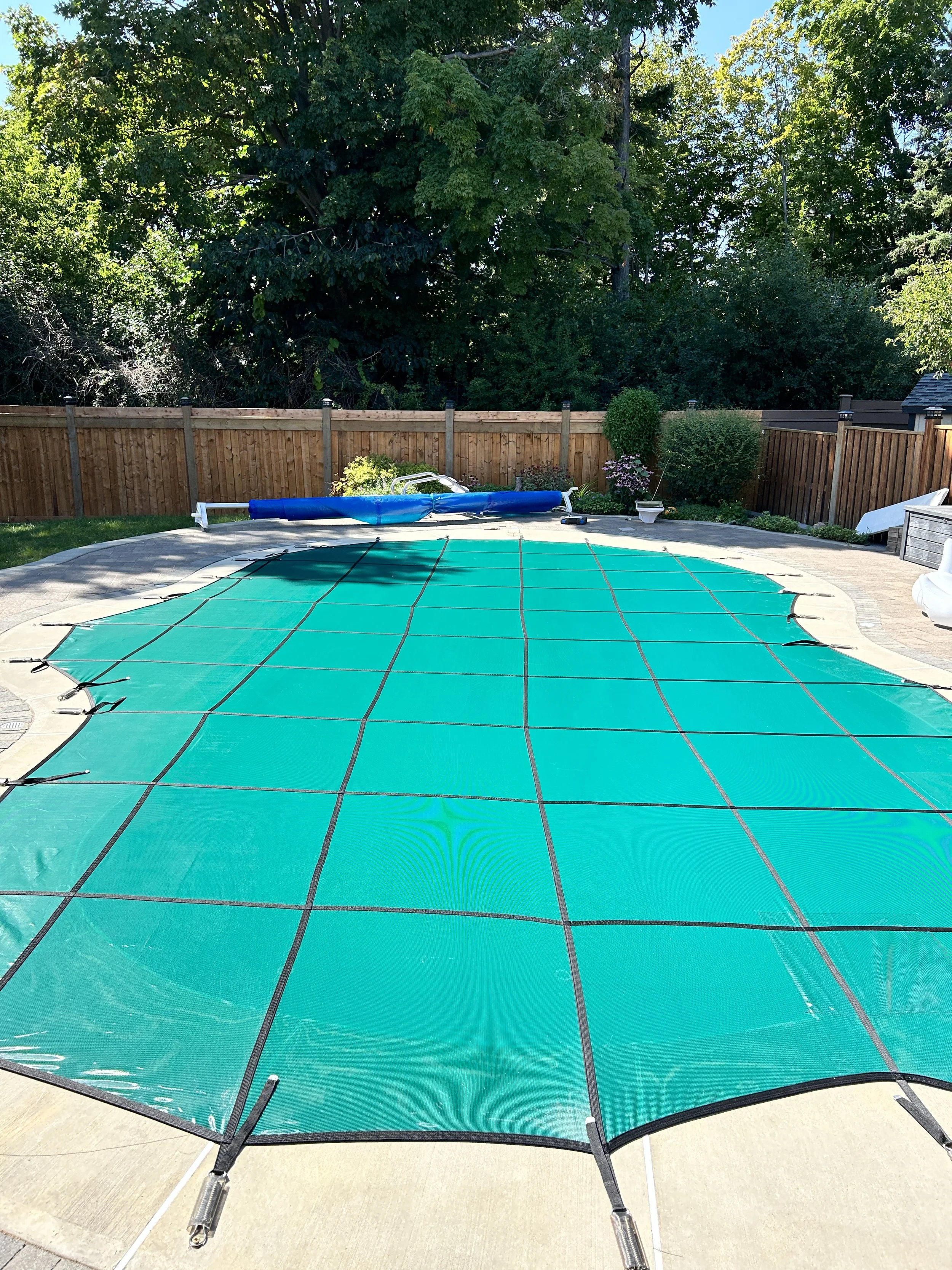 A swimming pool covered with a green pool cover, secured with black cables, in a backyard with a wooden fence and lush green trees.