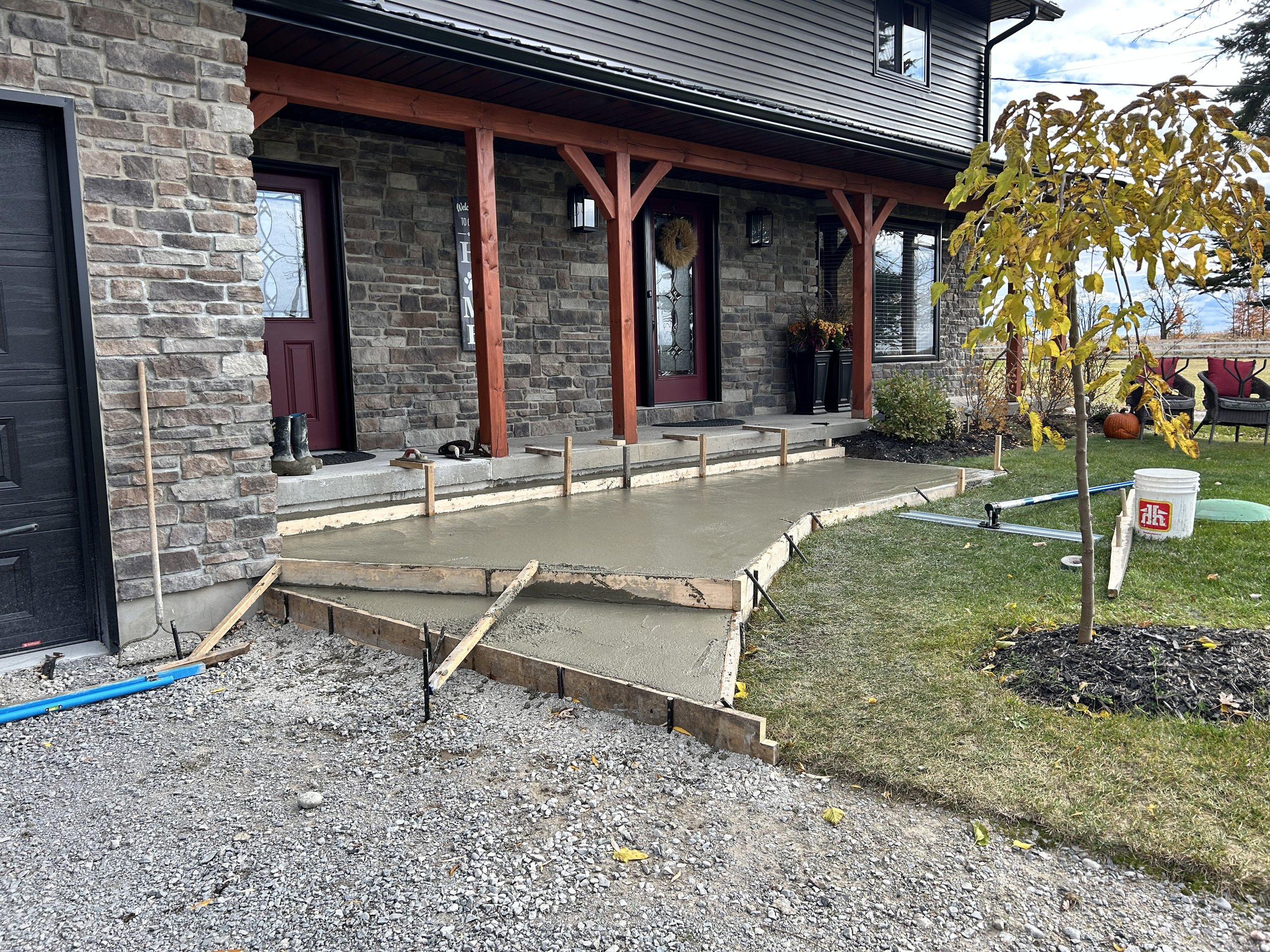 Concrete porch being poured in front of house with brick and siding exterior, steps leading to the entrance, and outdoor furniture and pumpkins on the grass.