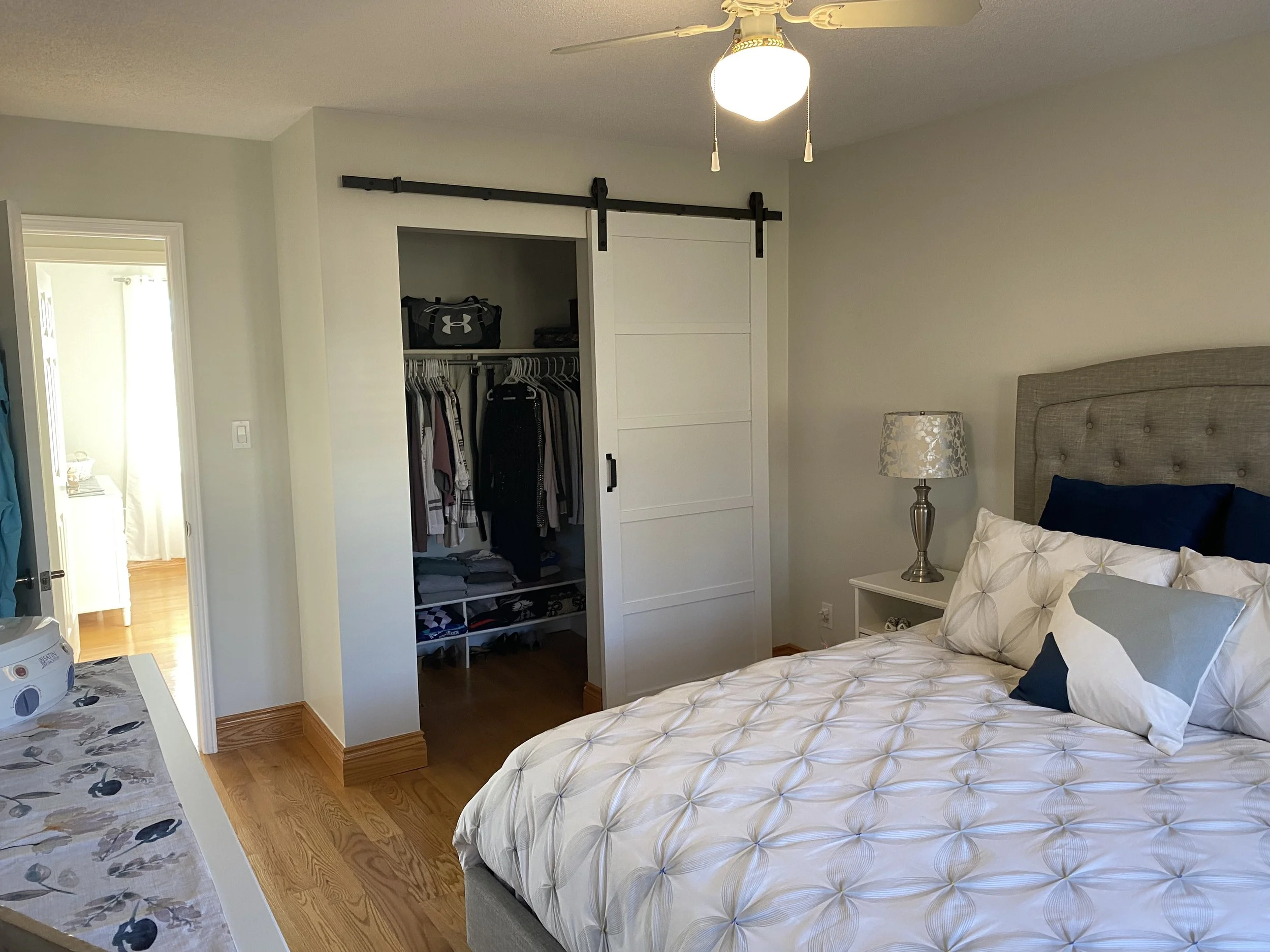 A bedroom with a bed, nightstand, lamp, open closet with hanging clothes, and a ceiling fan with light.