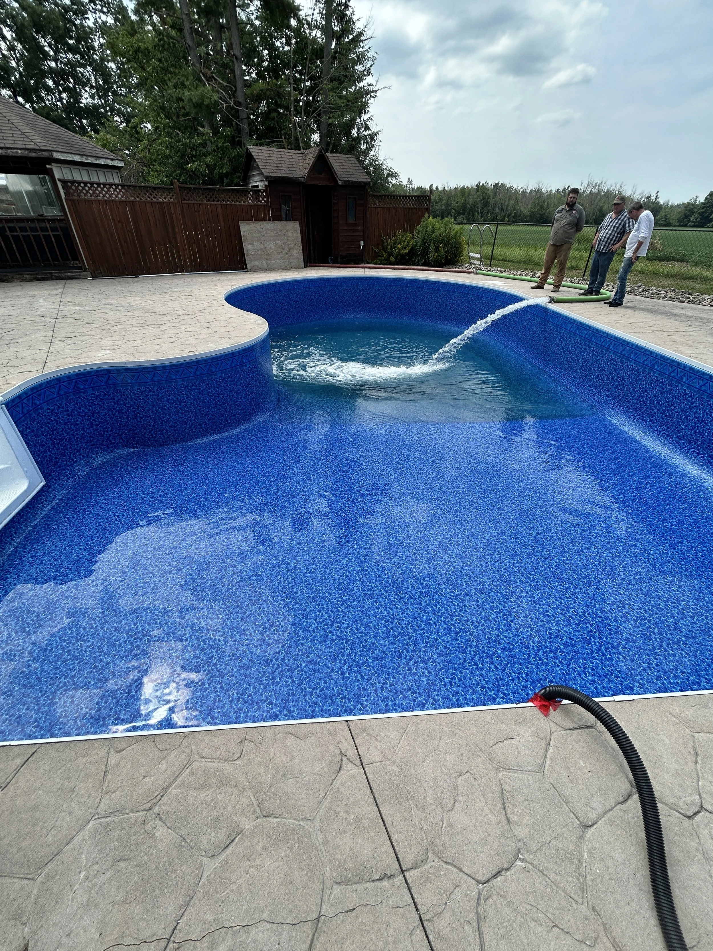 A newly installed in-ground swimming pool with blue lining, filled with water, on a concrete and stone patio. Three men are standing outside the pool near the edge, holding a hose that is filling the pool. In the background, there is a wooden fence, 