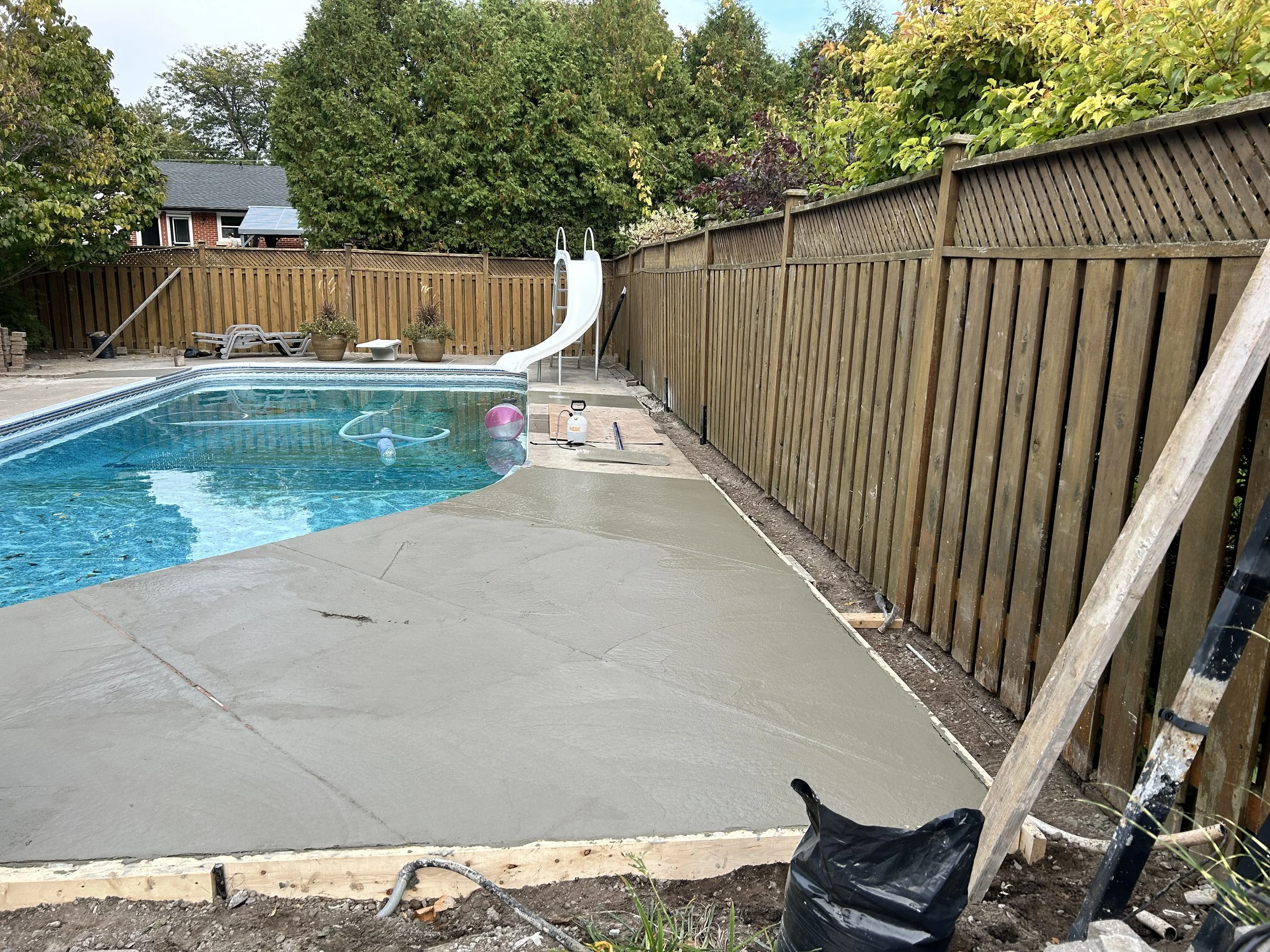 A backyard swimming pool under construction with a newly poured concrete deck, a slide, pool toys, and construction materials nearby.