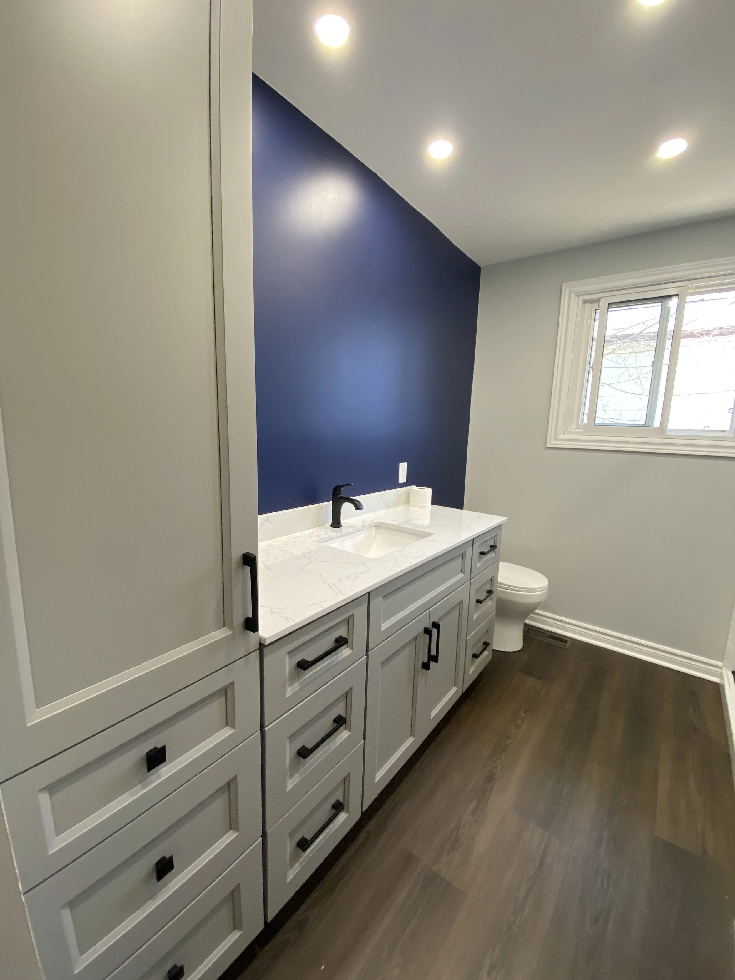 Modern bathroom with a blue accent wall, white vanity with black handles, marble countertop, black faucet, toilet, window, and wood flooring.