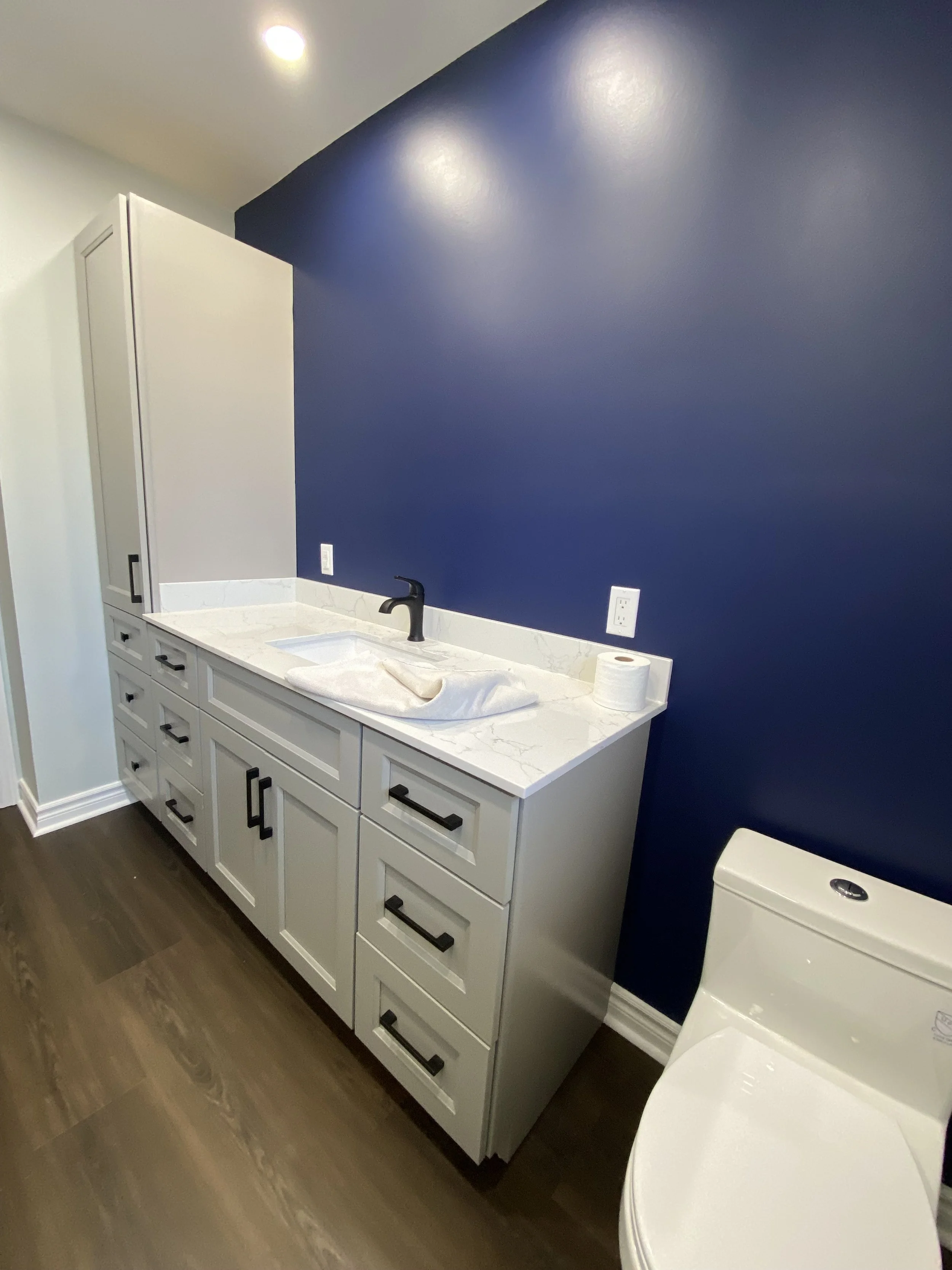 Bathroom vanity with white cabinet, marble countertop, black faucet, blue accent wall, toilet on the right, and hardwood flooring.