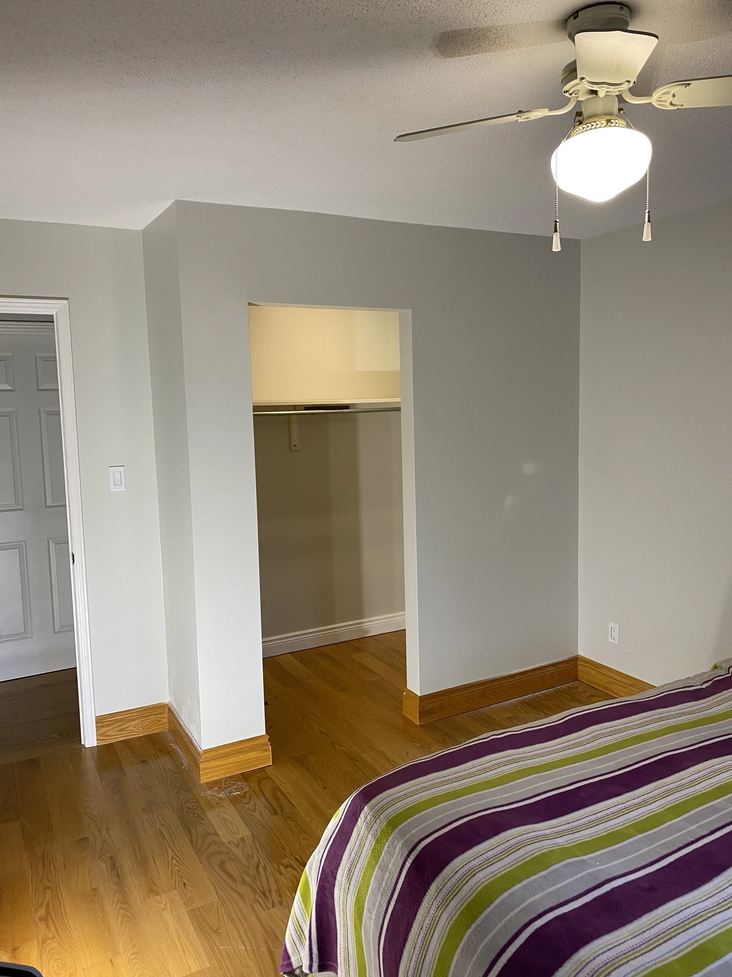 View of a bedroom corner with hardwood floor, light-colored walls, part of a bed with a striped purple, green, and white bedspread, an open closet with a single shelf, a ceiling fan with a light, and a door to another room or closet.