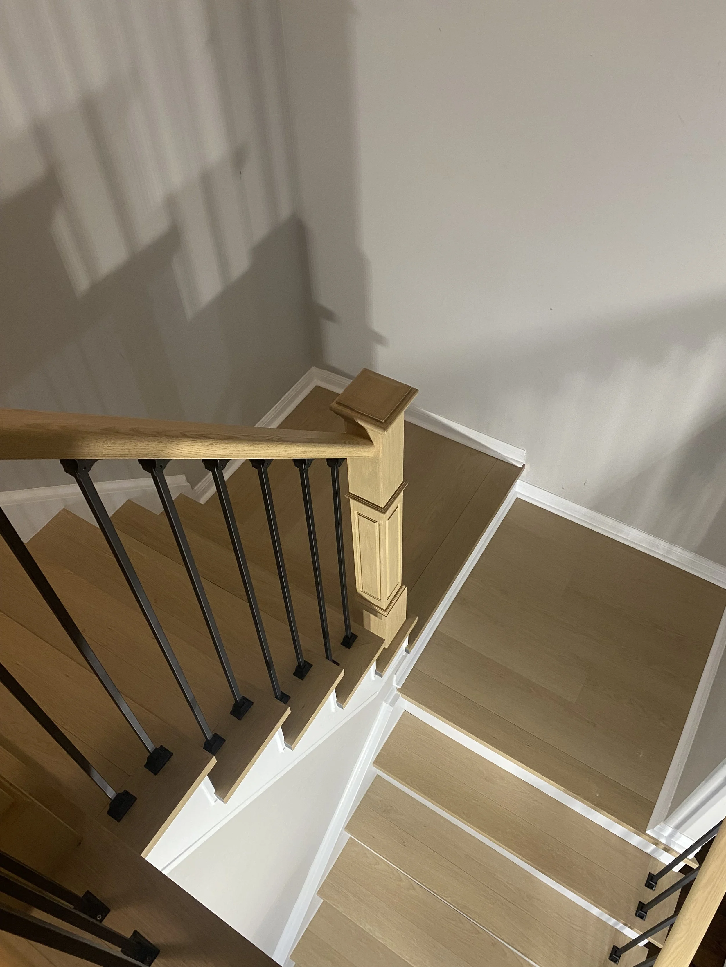 View of a staircase with wooden steps and black metal railing, showing the top of the staircase and the landing below, with a shadow cast on a white wall.