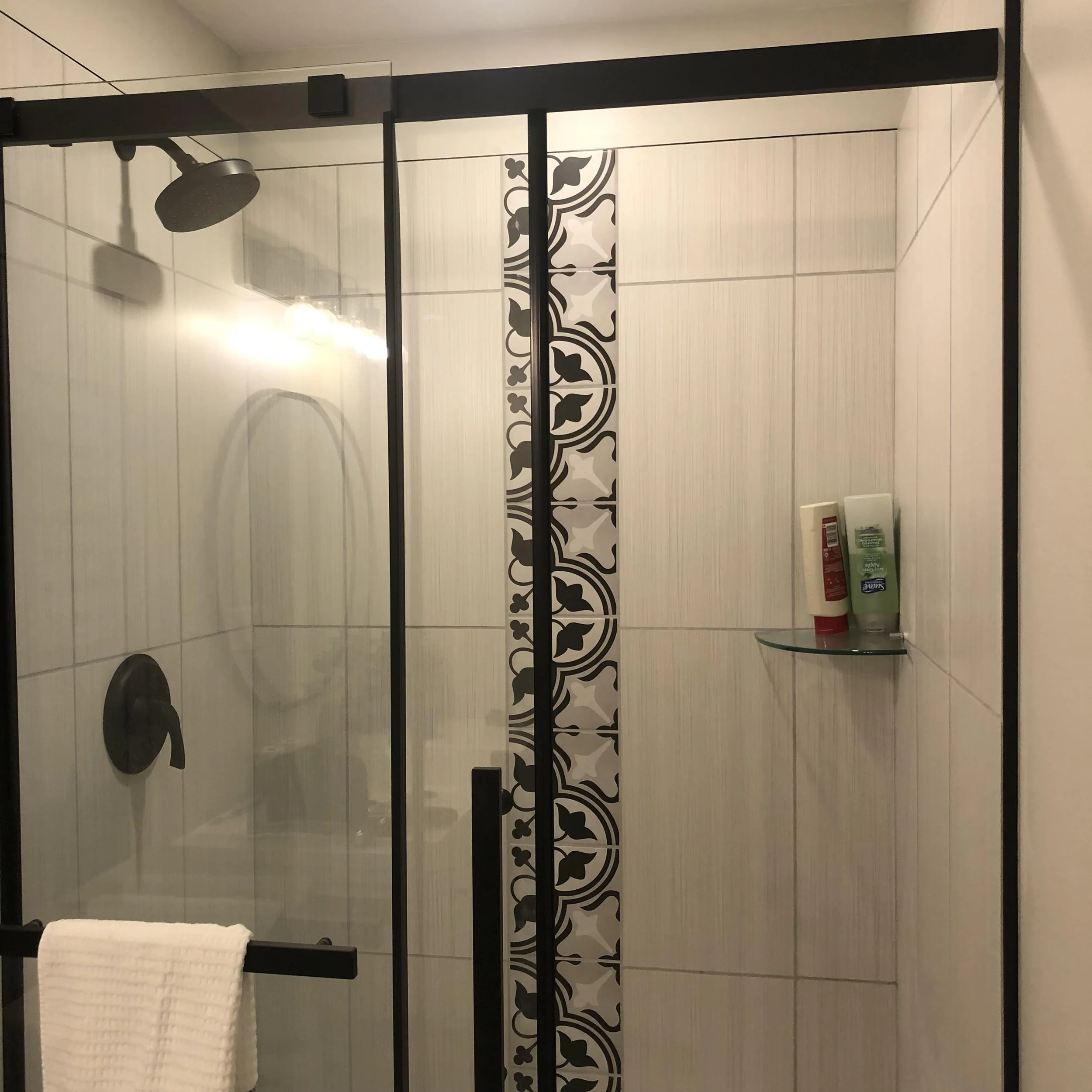 Inside a modern glass-enclosed shower with a black frame, decorative black and white tiles vertically aligned on the wall, a corner glass shelf with toiletries, a black showerhead, and a white towel hanging on a black towel bar.