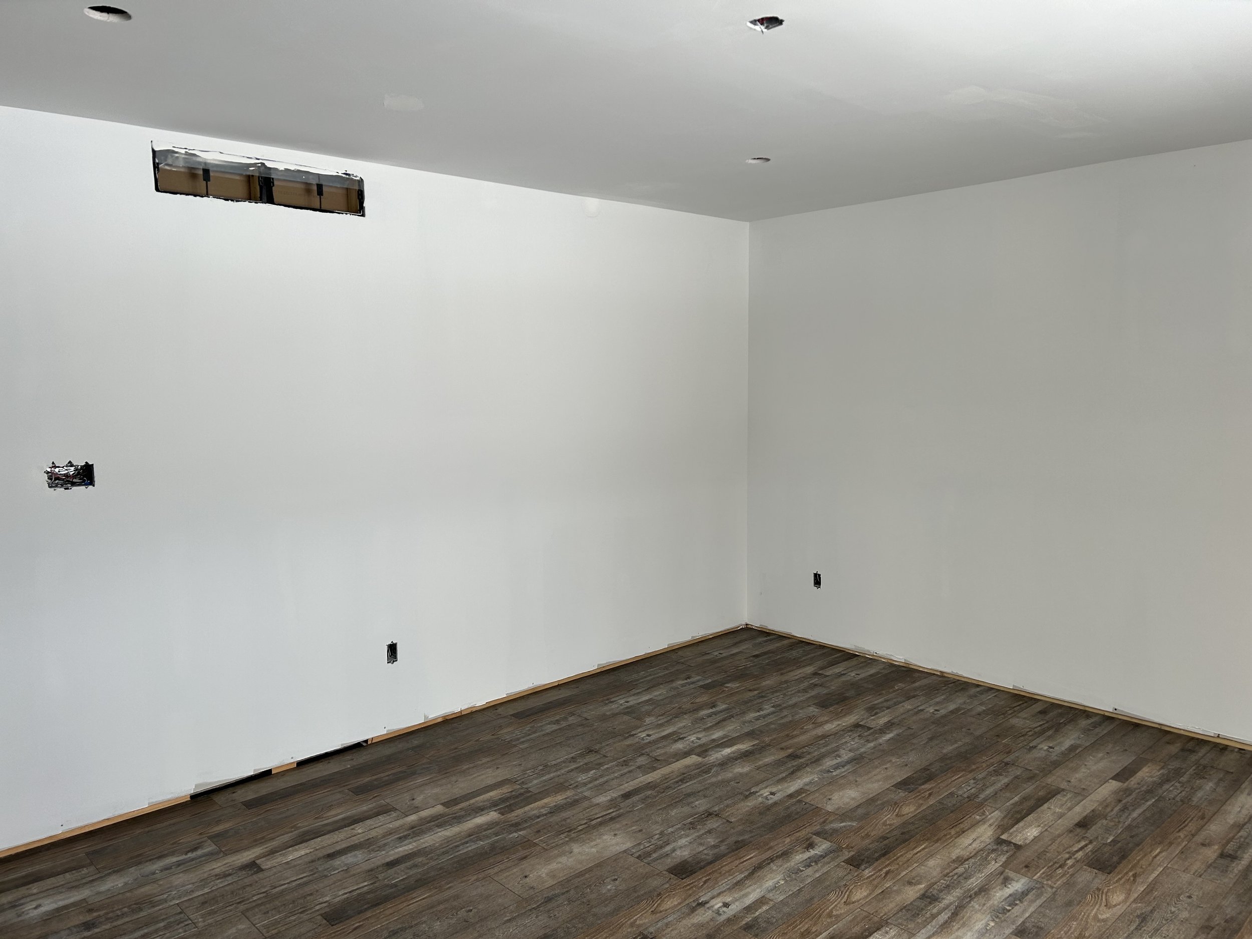 Empty room with white walls, hardwood floor, electrical outlets, and ceiling fixtures under construction.