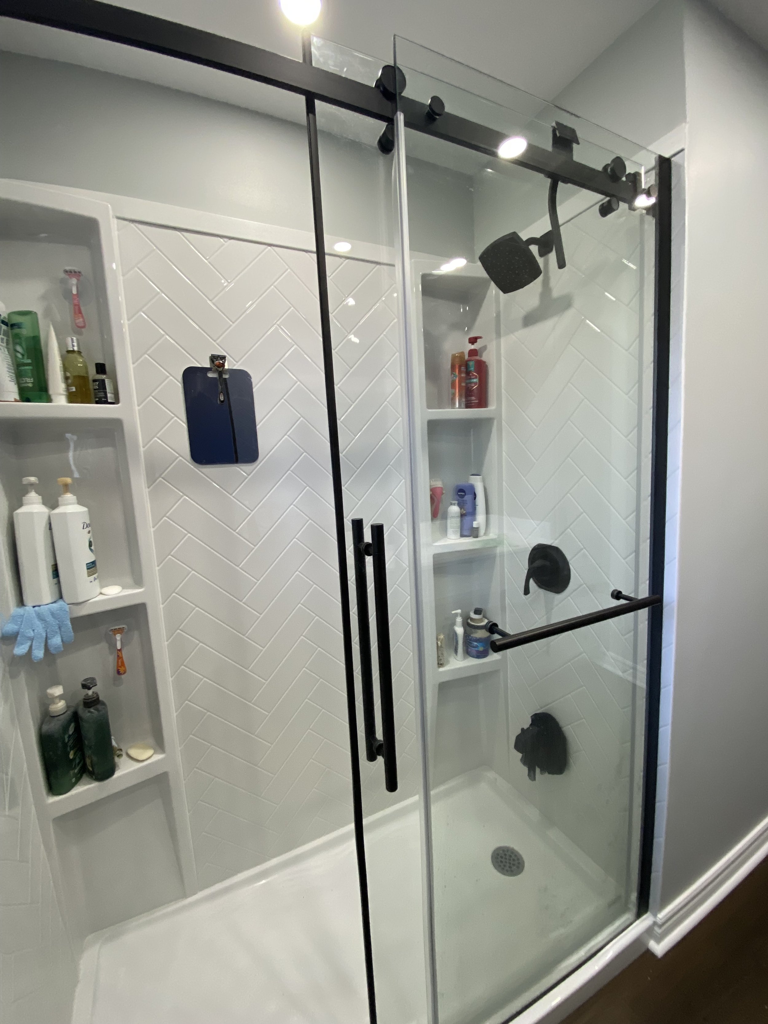 A modern glass-enclosed shower with black fixtures, white tiled walls, and built-in shelves holding toiletries.