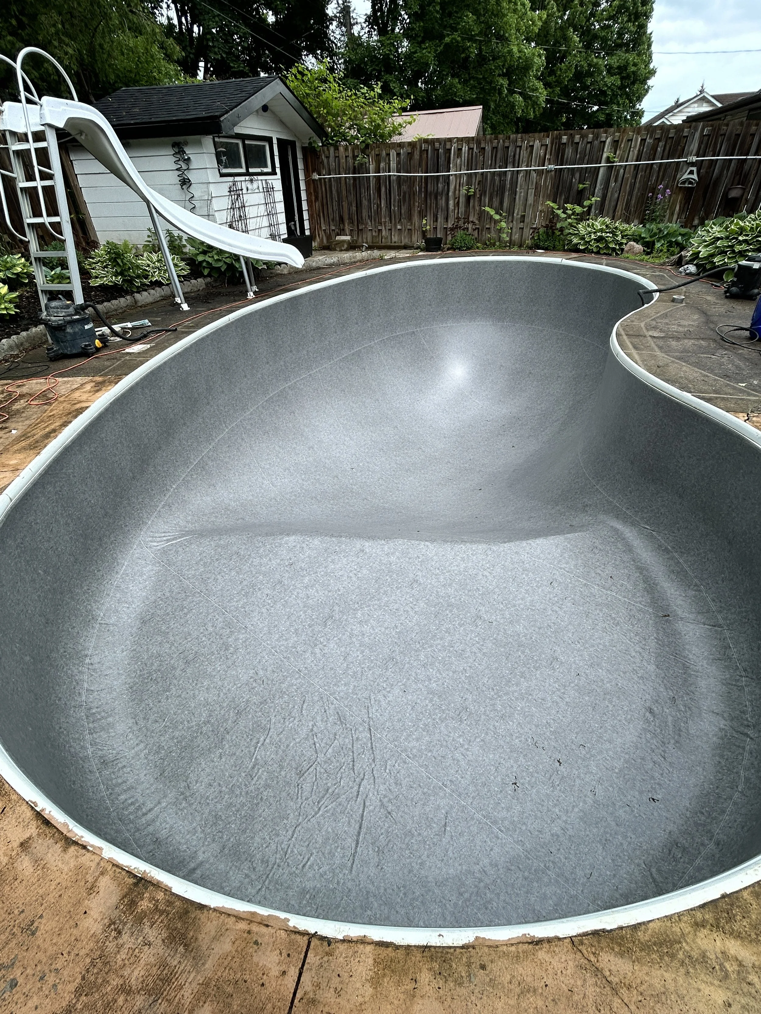 Empty backyard swimming pool under construction, with a slide and garden in the background.
