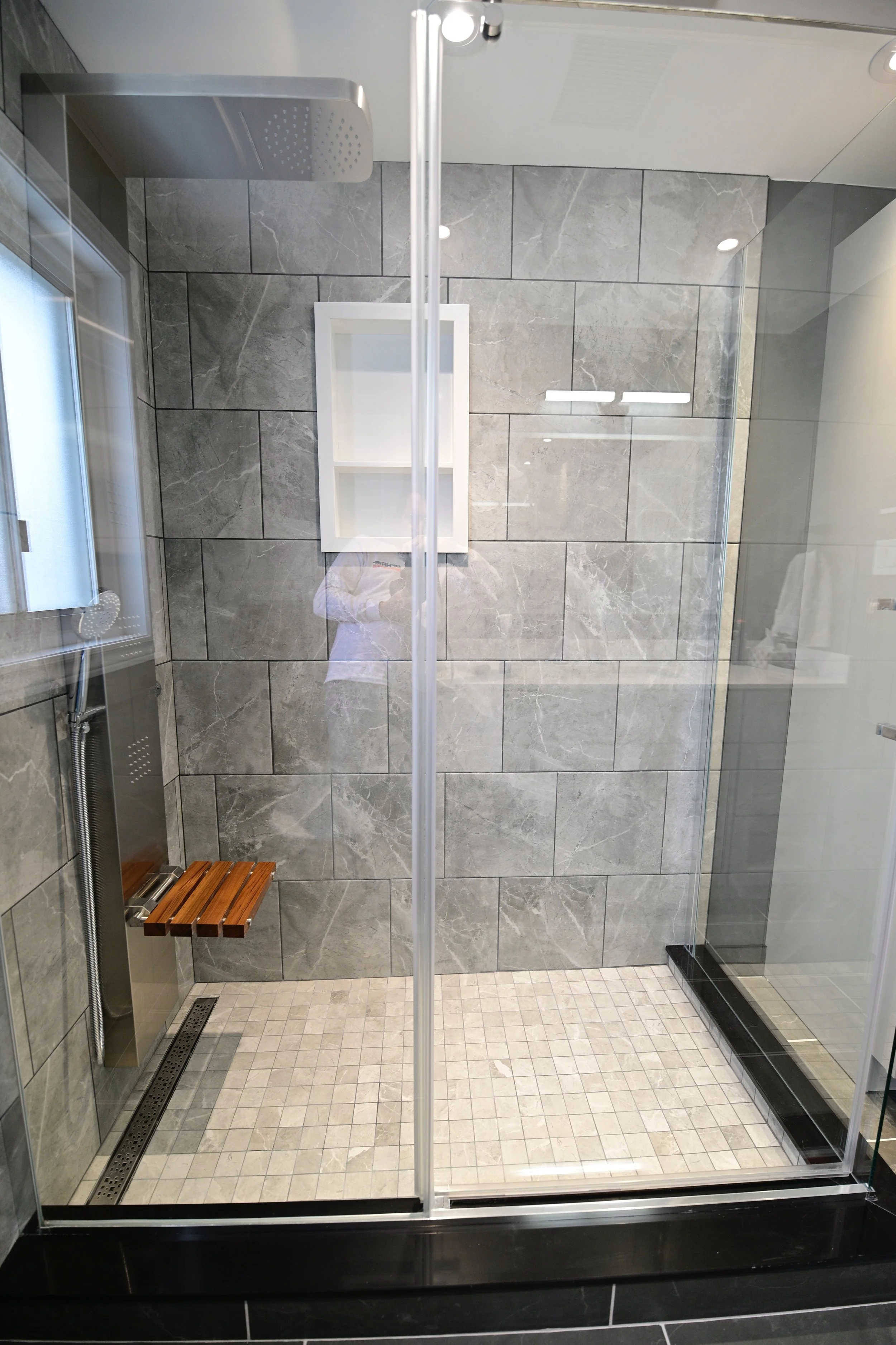 Modern walk-in shower with gray marble tile walls, small beige tiled floor, a wooden bench, a glass door, a niche shelf, and a rainfall shower head.