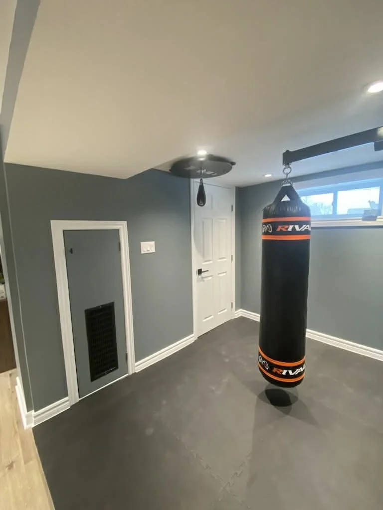 A basement room with gray walls and a dark gray floor, featuring a hanging punching bag near a small window and a closed white door.