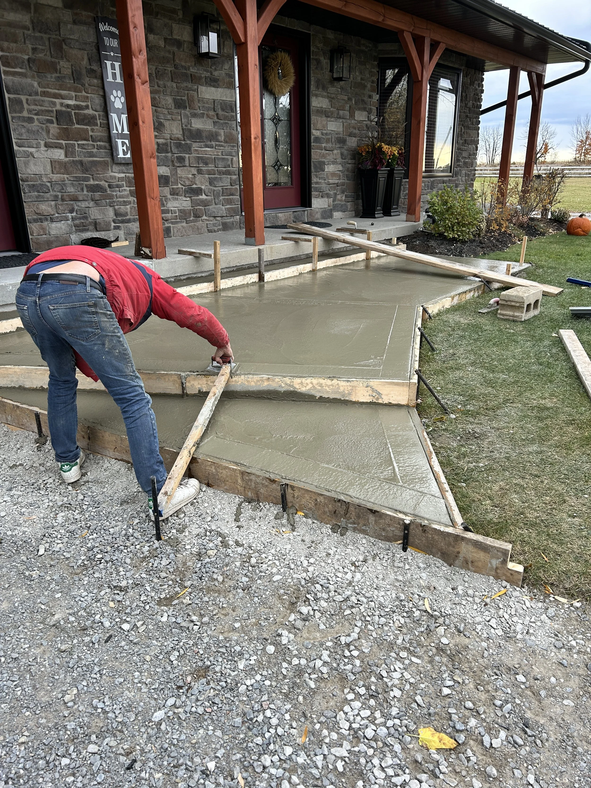 A person in a red shirt and jeans is leveling wet concrete for a new sidewalk or porch in front of a house with a covered porch entrance. The house has brick walls, a decorative door, hanging lantern lights, and flower arrangements. The construction 