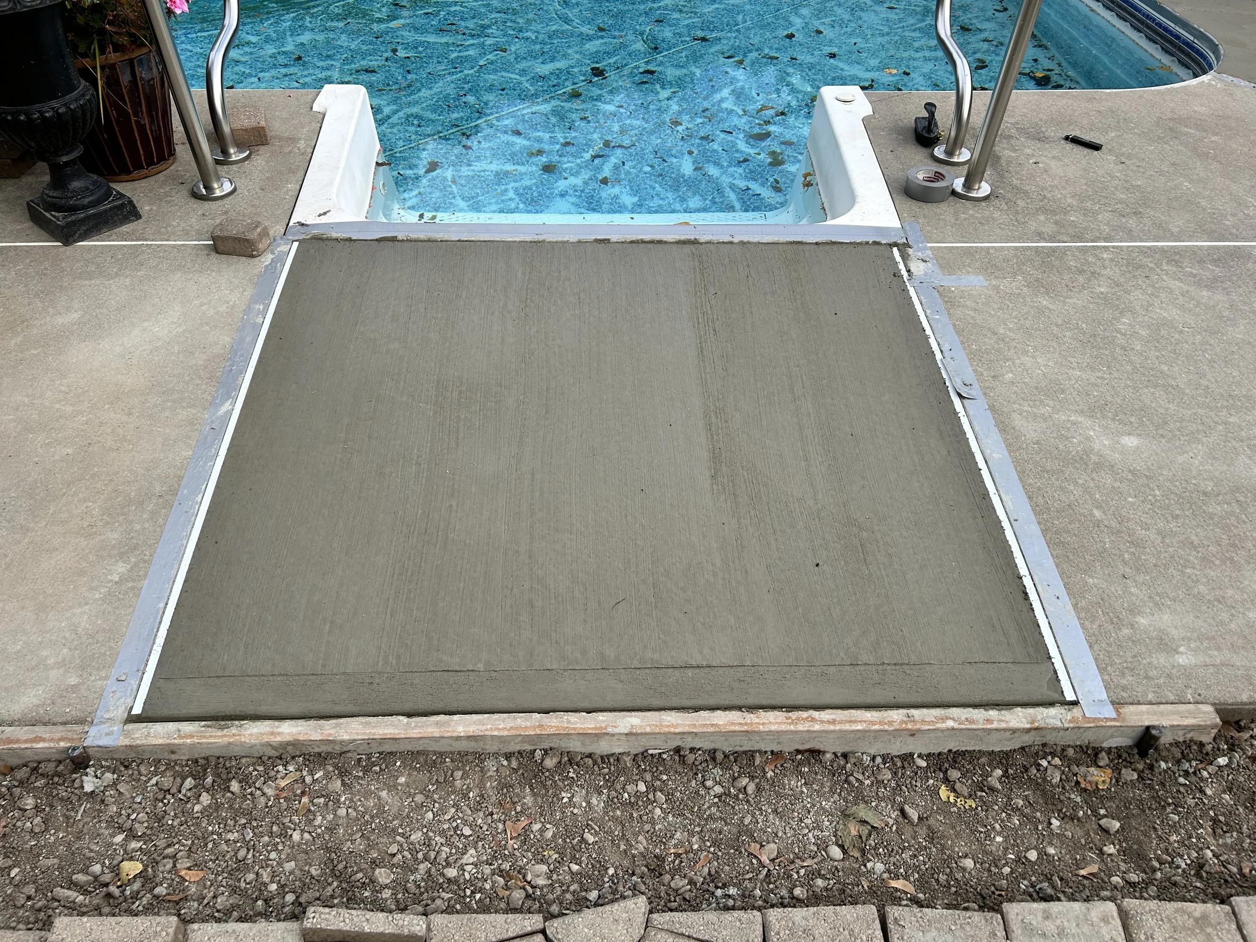 A freshly paved concrete area near a swimming pool, with metal handrails and tools visible around.