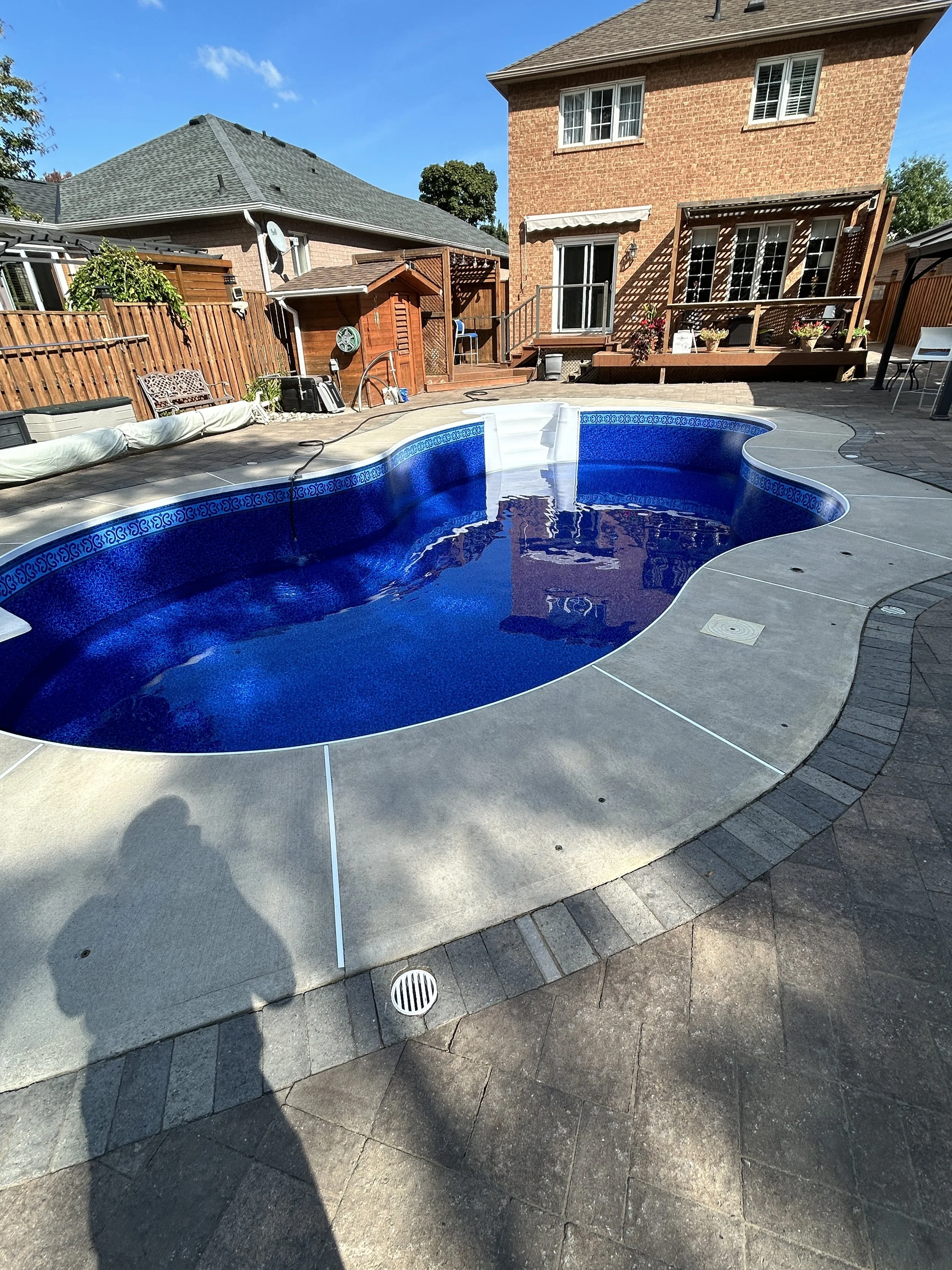 Backyard with in-ground swimming pool, surrounded by a concrete and brick patio, with a wooden fence, a brick house with a deck and garden next to it, and a clear blue sky.