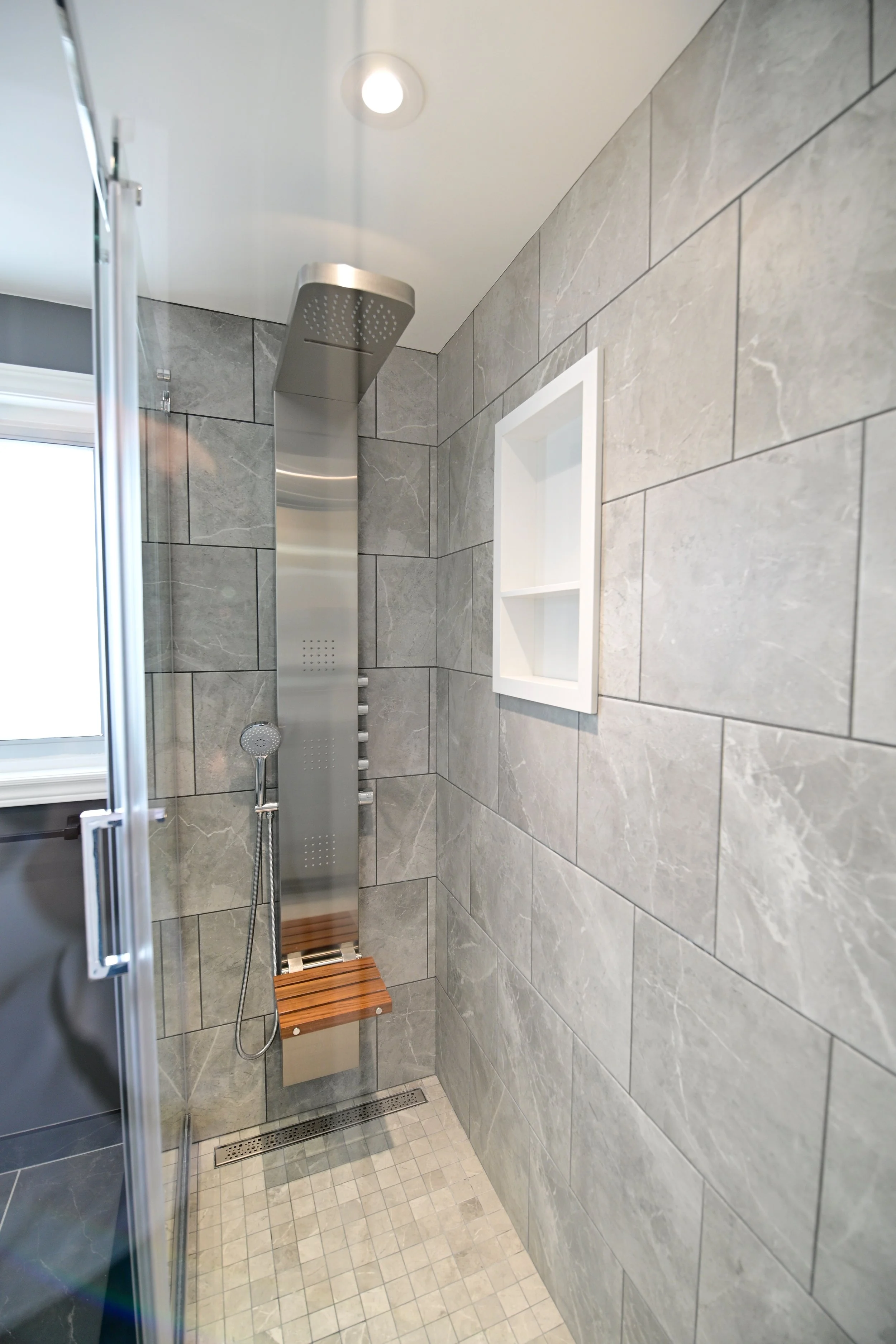 Modern walk-in shower with gray tiled walls, a stainless steel shower panel with a handheld shower spray, a small built-in wall niche, and a window with a white frame.