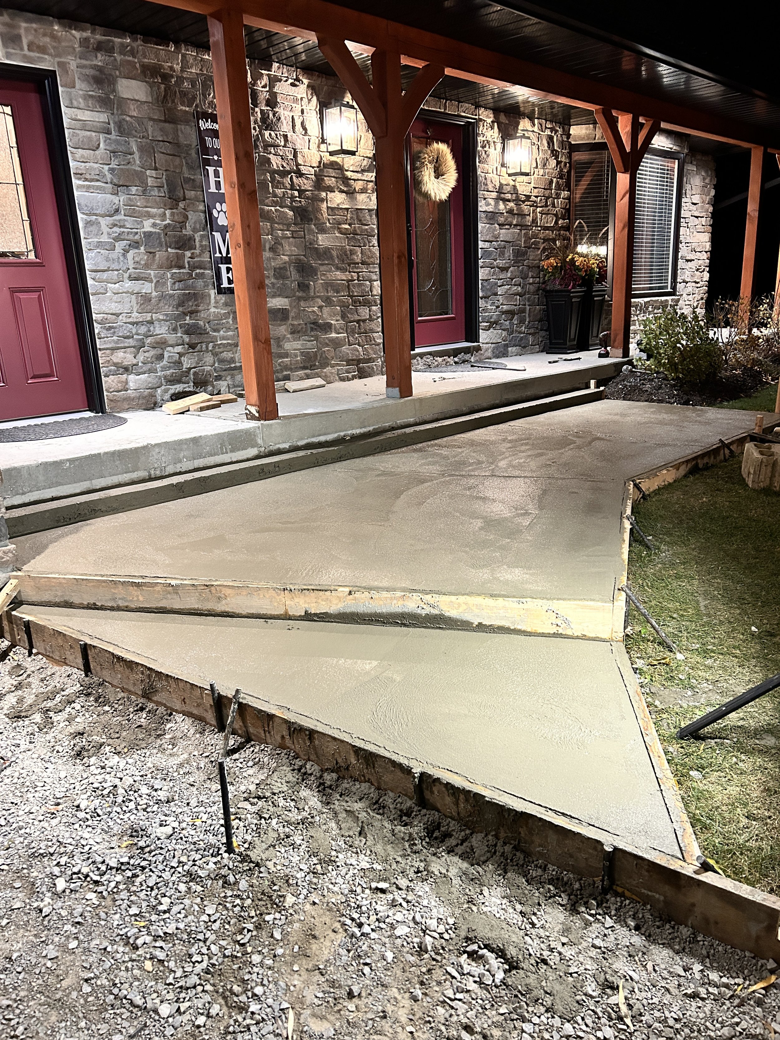 Concrete being freshly poured and smoothed on a walkway in front of a house with stone exterior walls, maroon front doors, and porch lighting.